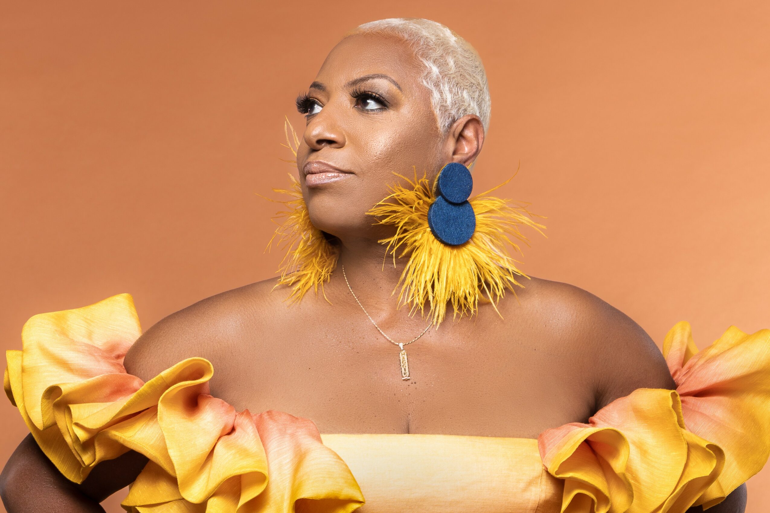 Woman with short platinum blonde hair wearing large yellow feather earrings and ruffled yellow top against a plain background.