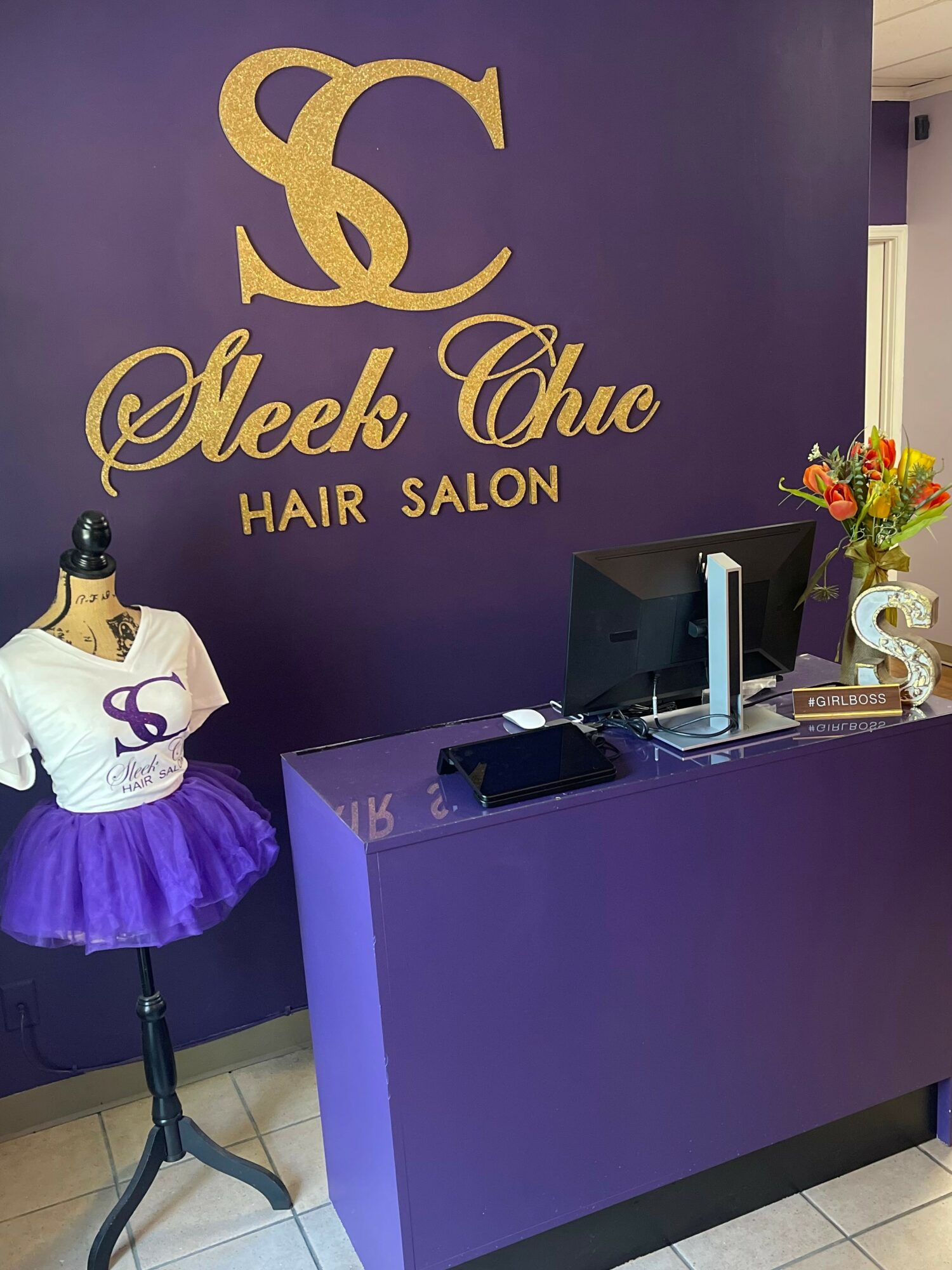 Reception desk with computer, flowers, and a mannequin wearing a purple skirt and white top, in front of a purple wall with gold logo and text.