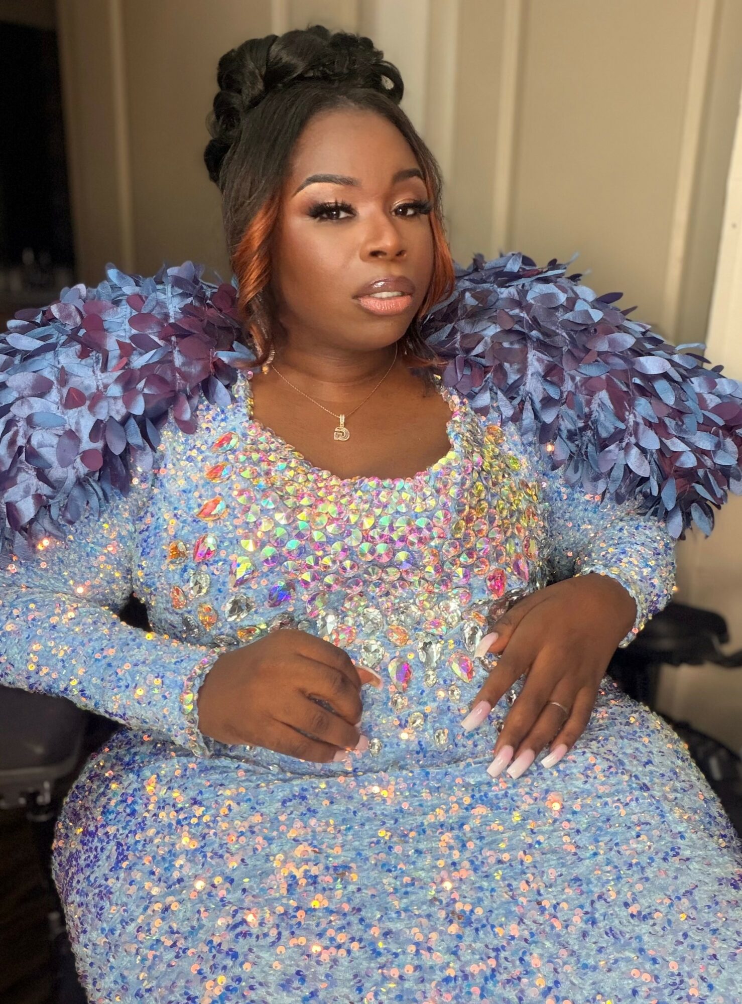 Woman in a colorful, sparkly dress with large purple feathered shoulders, sitting indoors with a neutral background.