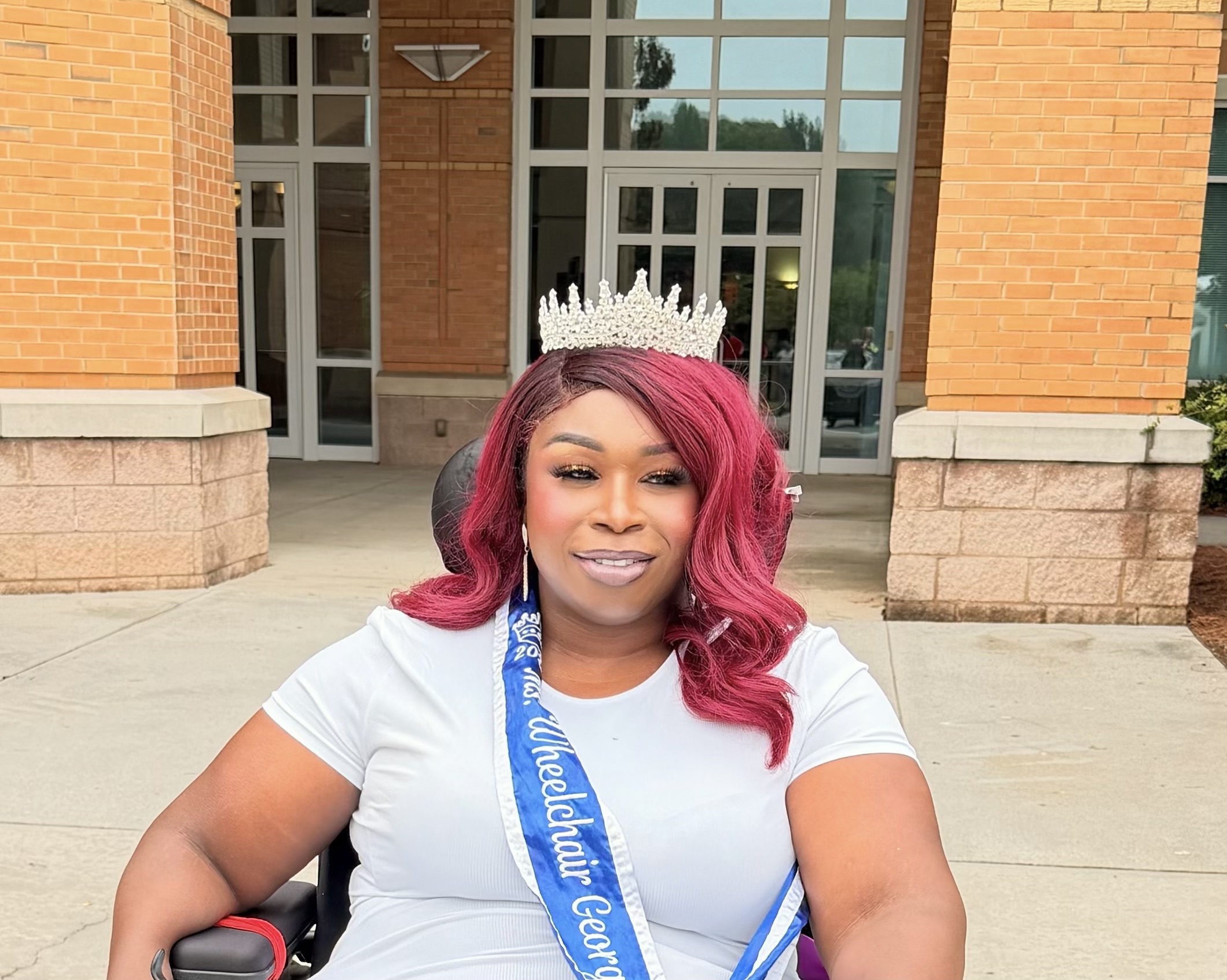 Woman with pink and black hair wearing a crown and sash, smiling outdoors in front of a brick building.