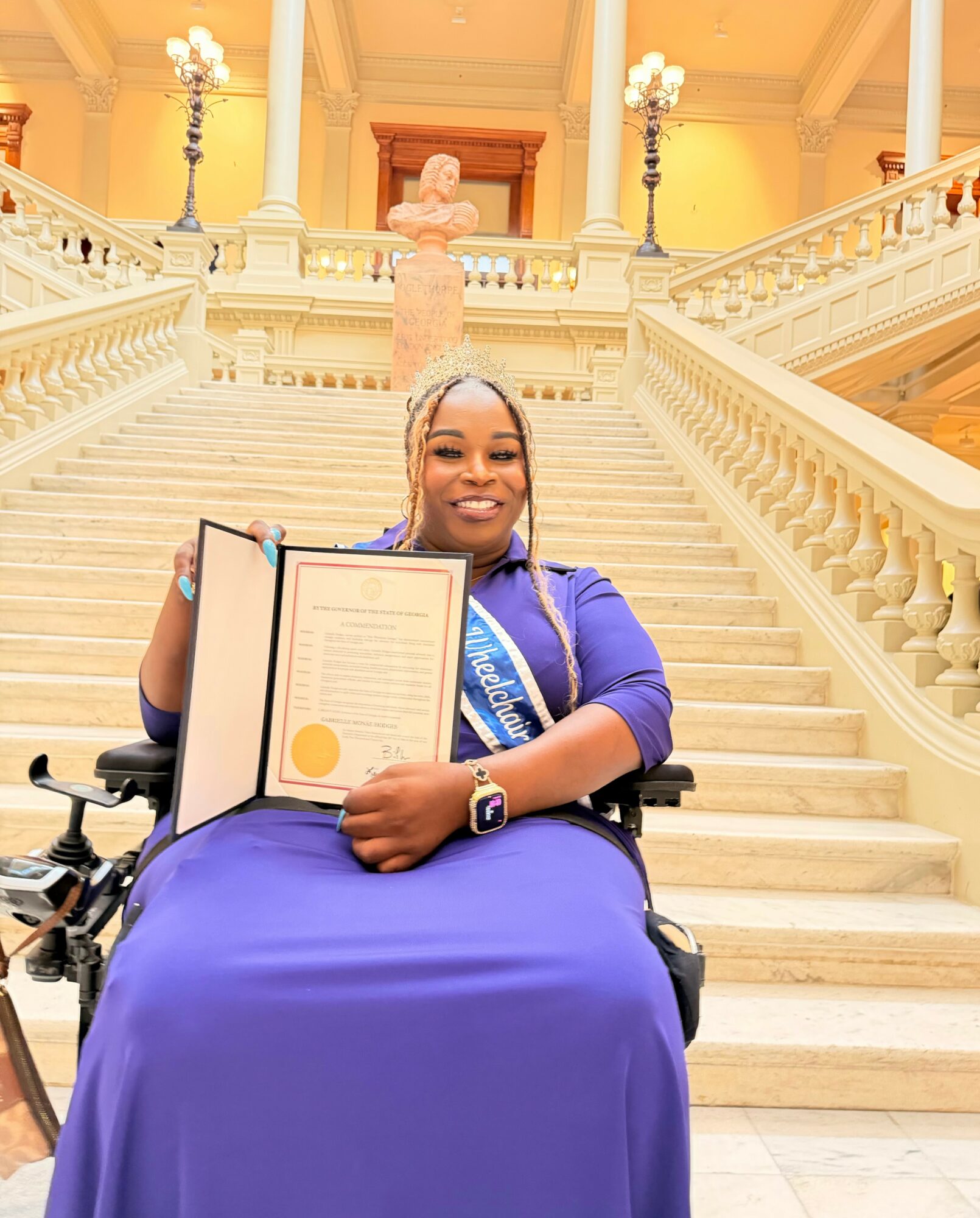 Young woman in a wheelchair holding a diploma, smiling, in a grand staircase hall with ornate decor.