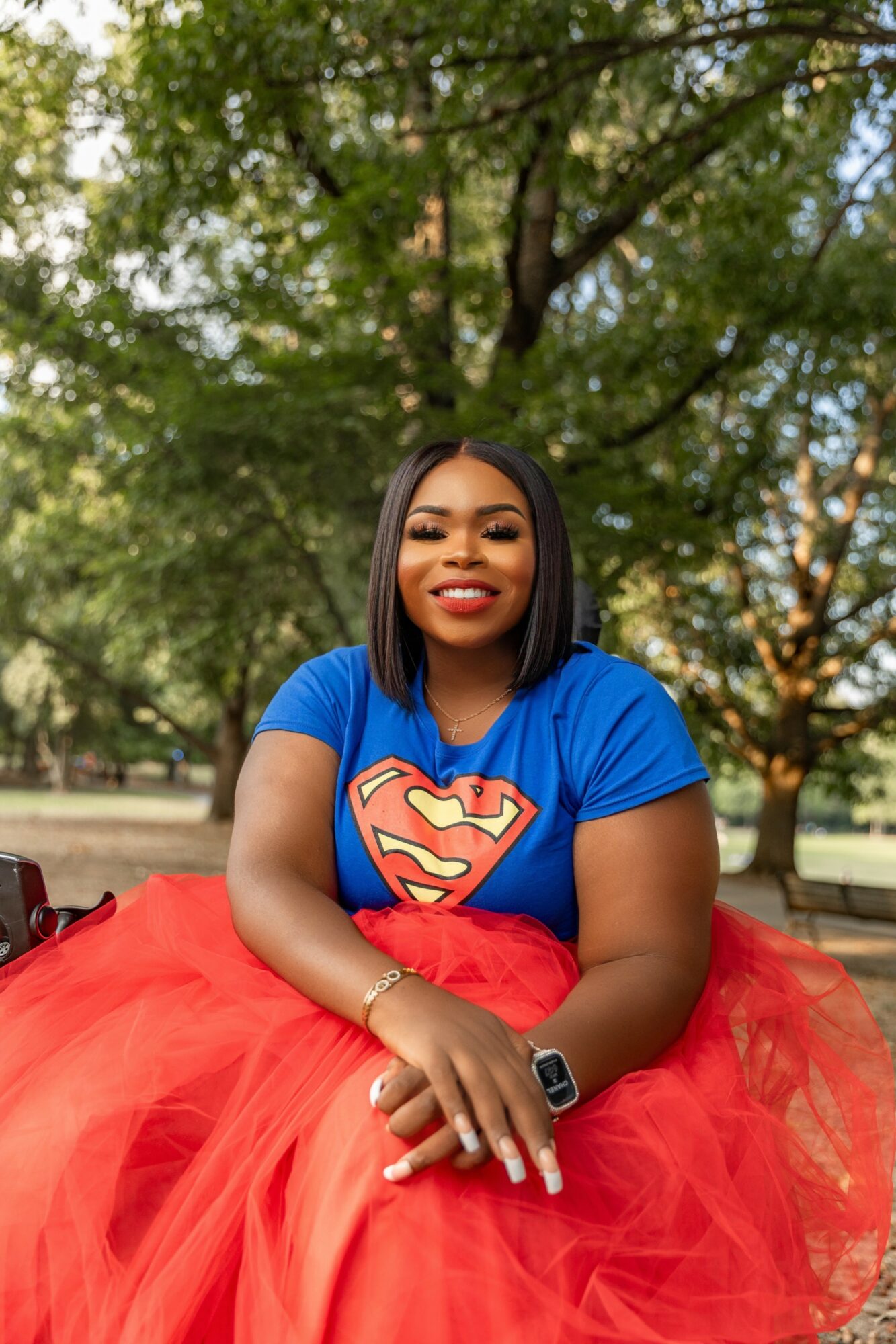 Young woman with shoulder-length black hair, wearing a blue Superman shirt and red tulle skirt, smiling outdoors with trees in background.