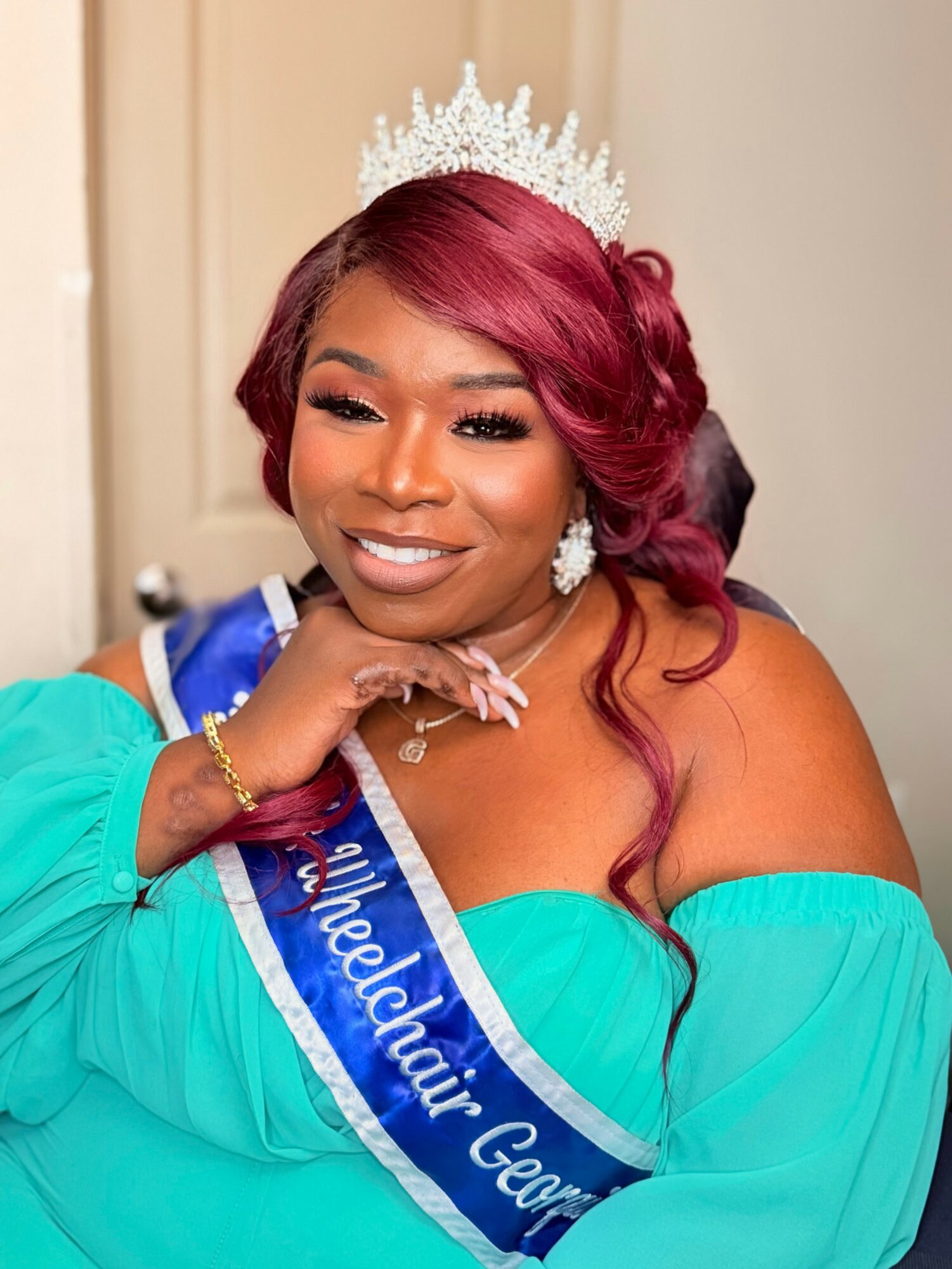 Woman wearing a crown and sash, smiling, with long wavy hair, in a turquoise dress, indoors.
