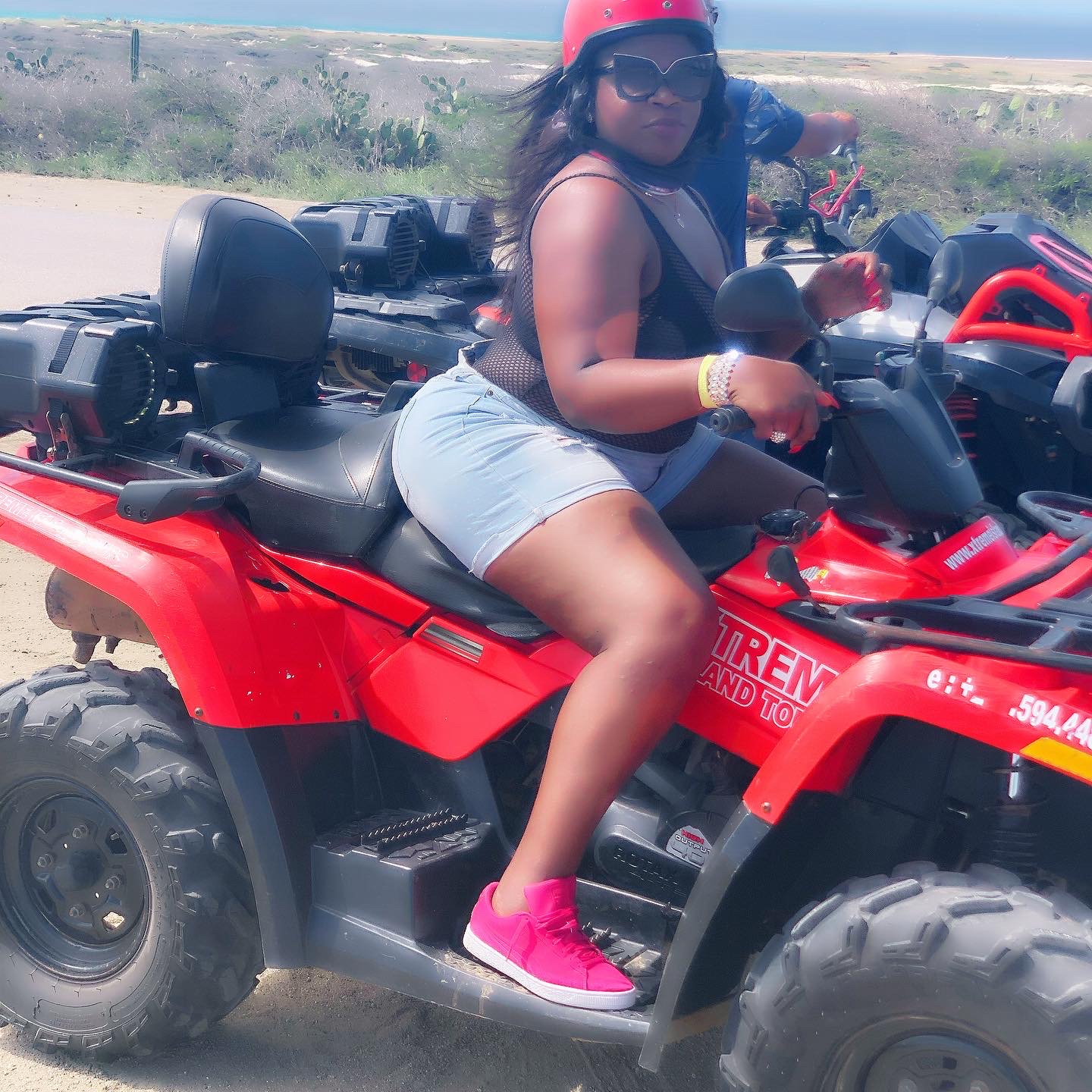 Person sitting on a red all-terrain vehicle outdoors, wearing a helmet, sunglasses, and pink shoes, with a sandy background.