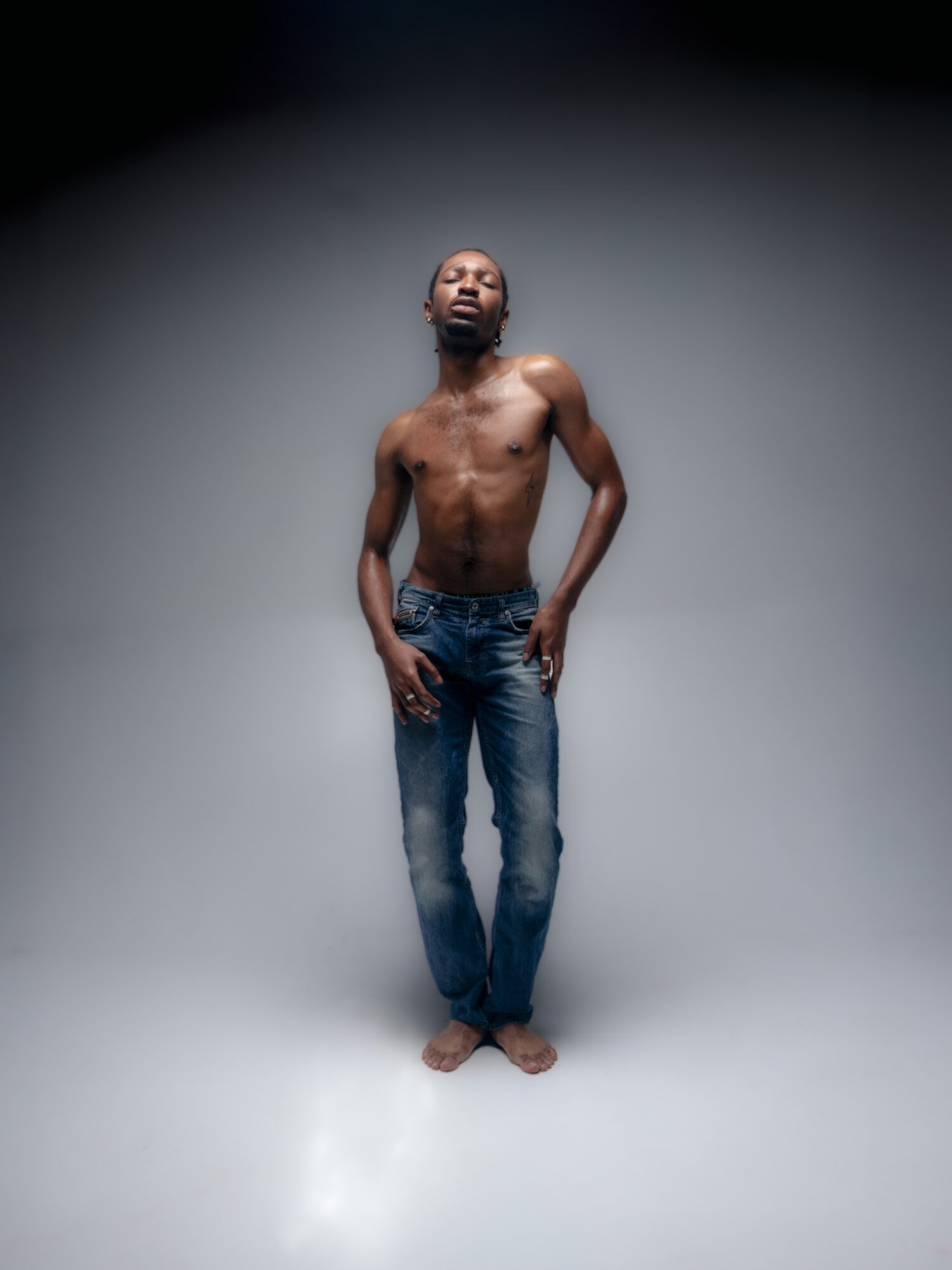 A shirtless man stands with hands in pockets against a plain background.