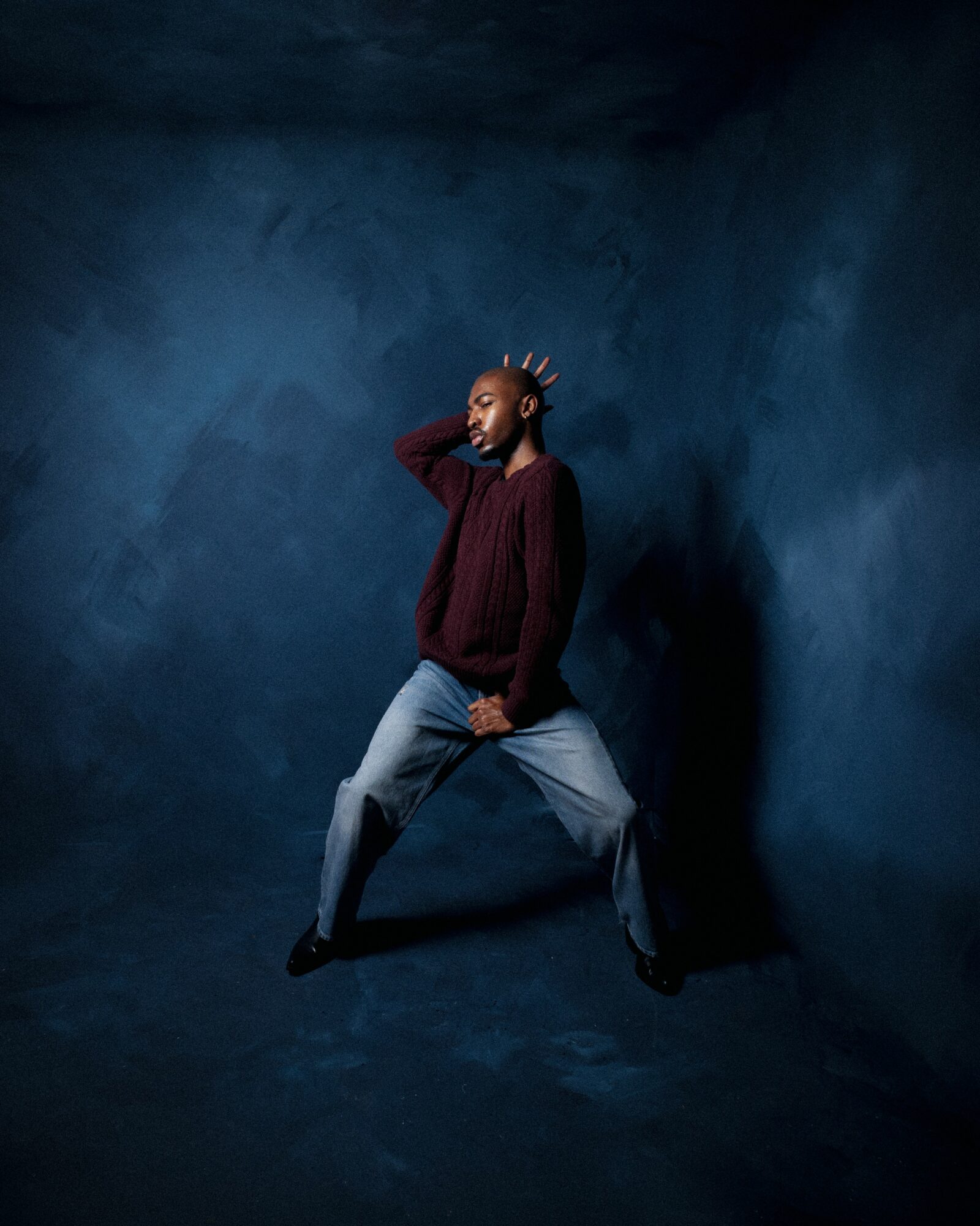 Person with braided hair in a maroon sweater and gray pants stands against a dark blue textured wall.
