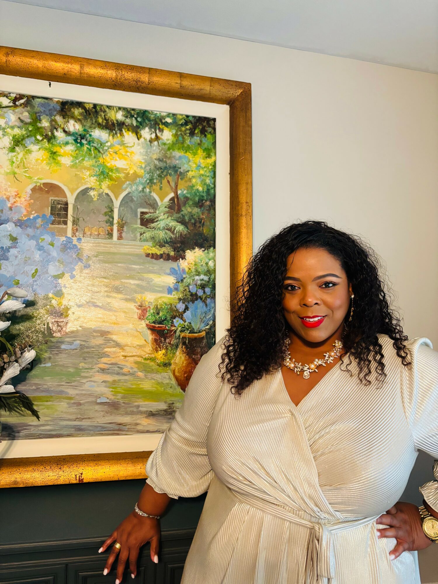 Woman with curly black hair and red lipstick standing next to a framed landscape painting, wearing a white dress and pearl necklace.