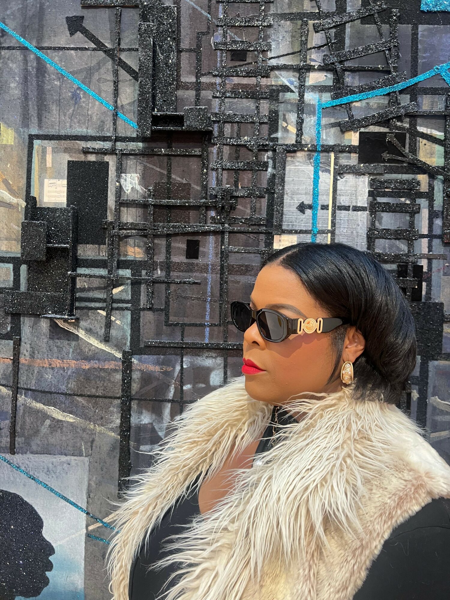 Woman with black hair, sunglasses, red lipstick, wearing a fur vest, standing outdoors with a city background.