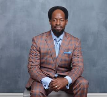Man in a plaid suit sitting against a gray background, hands clasped, looking at the camera.