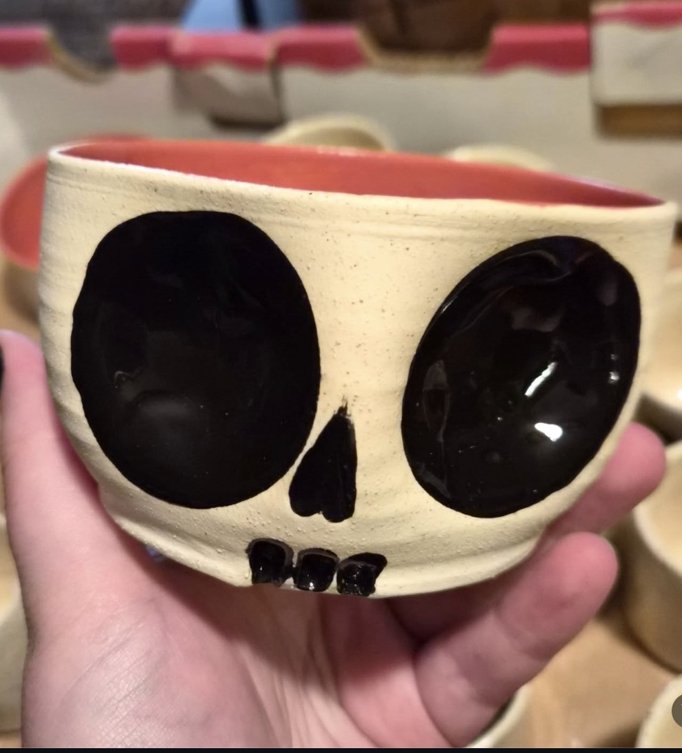 Hand holding a ceramic bowl decorated as a skull with black eye sockets and nose, and teeth, against a blurred background.