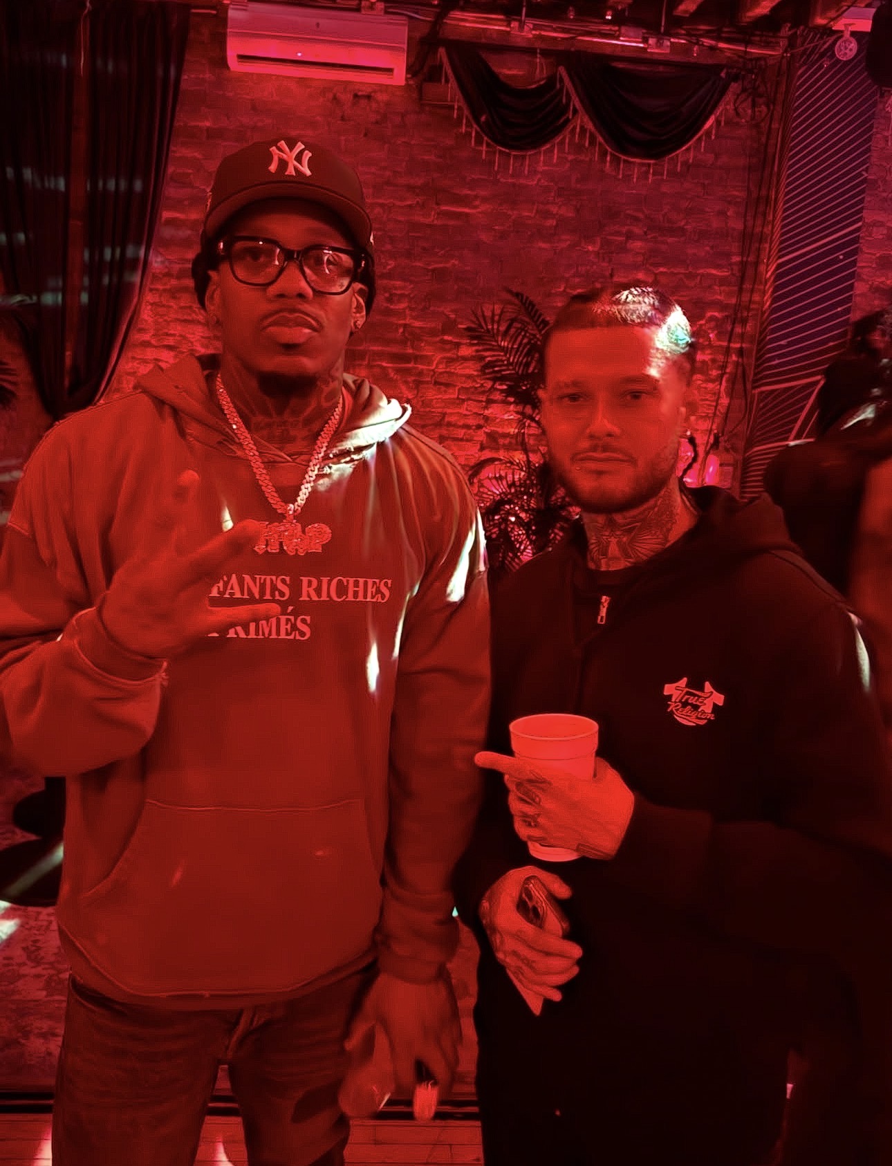 Two men standing indoors, one holding a cup, with a brick wall and curtains in the background, illuminated by red lighting.