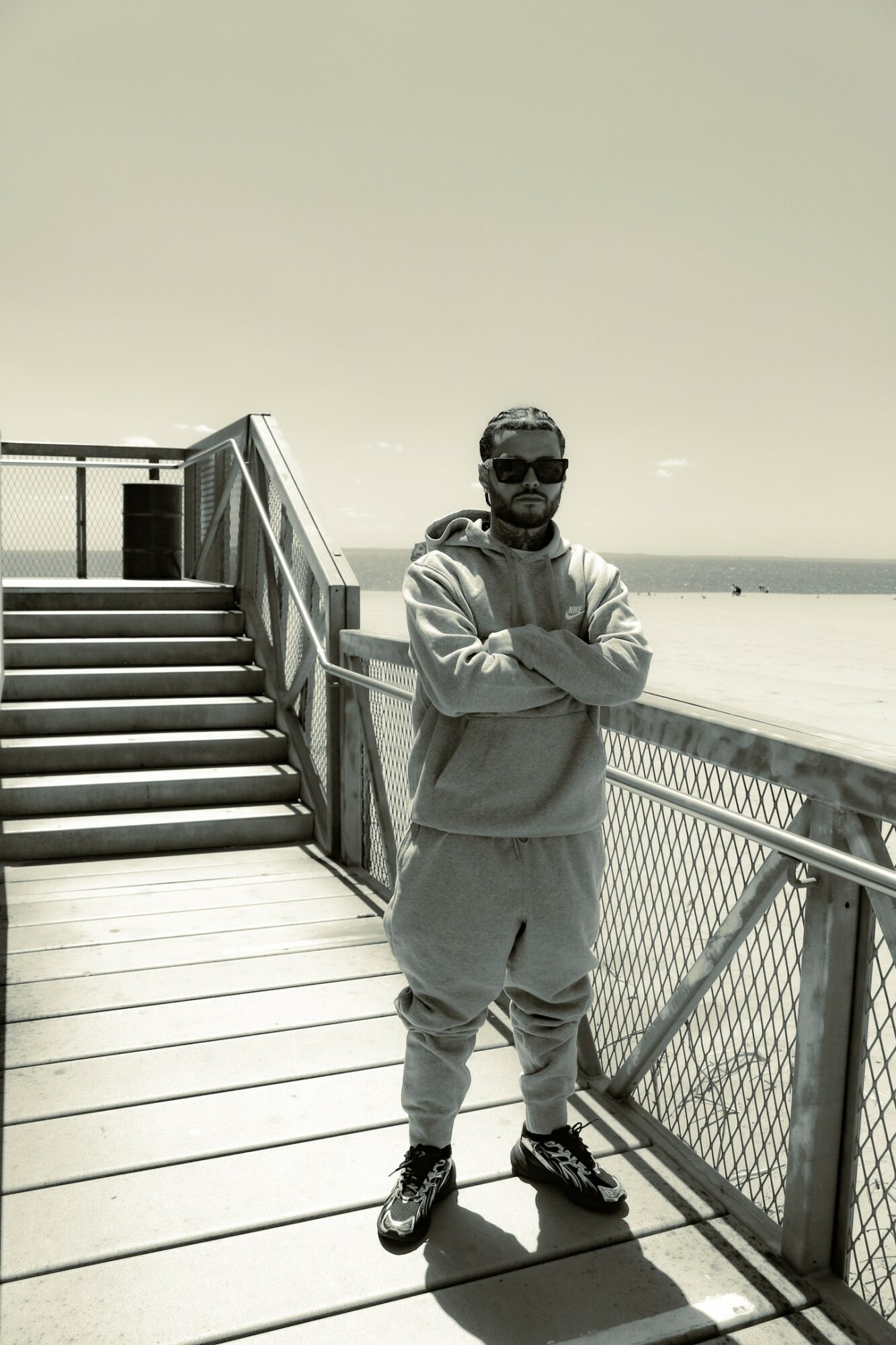 Man with sunglasses and beard stands with arms crossed on a boardwalk near the beach, railing behind him, sky above.