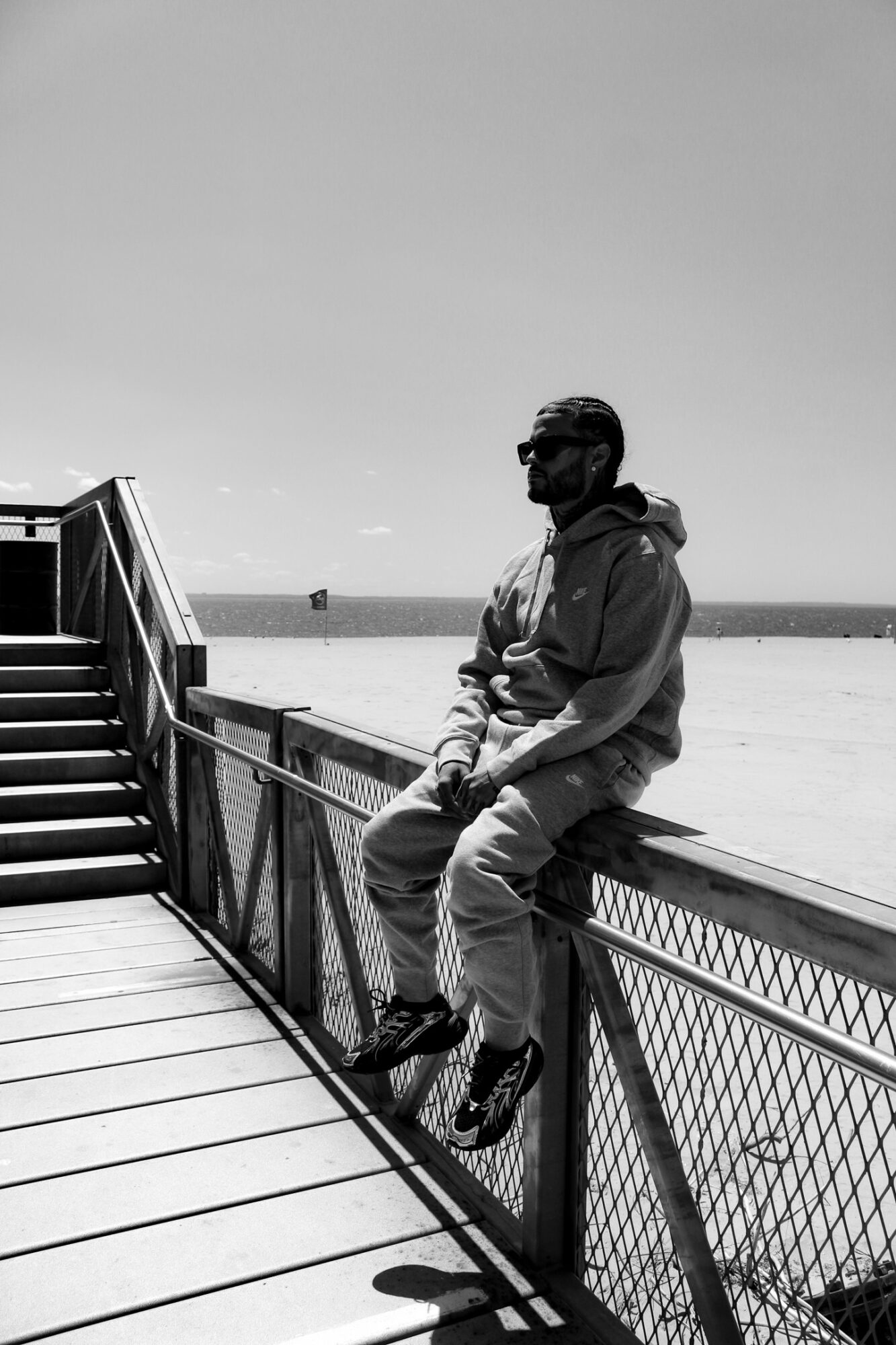 Person sitting on a railing near a beach, wearing sunglasses and a jacket, with a clear sky and ocean in background.