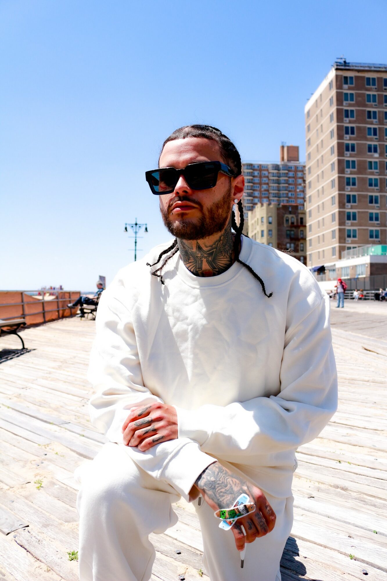 Man with sunglasses and tattoos, wearing white outfit, standing outdoors on a sunny day with buildings in background.