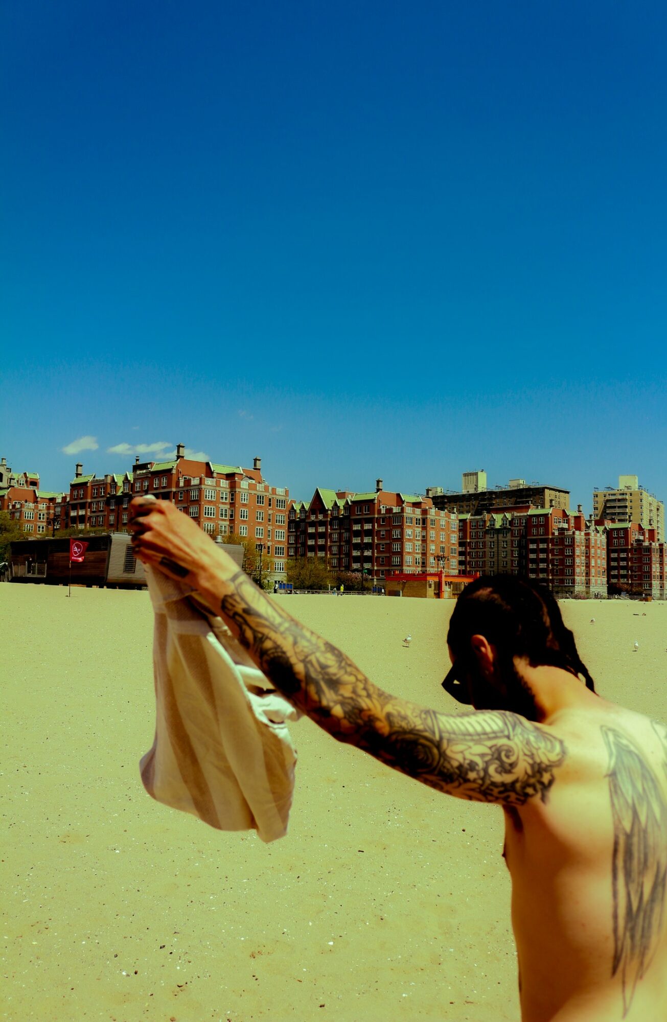 Person with long tattooed arm holding a bag against a city skyline and blue sky.