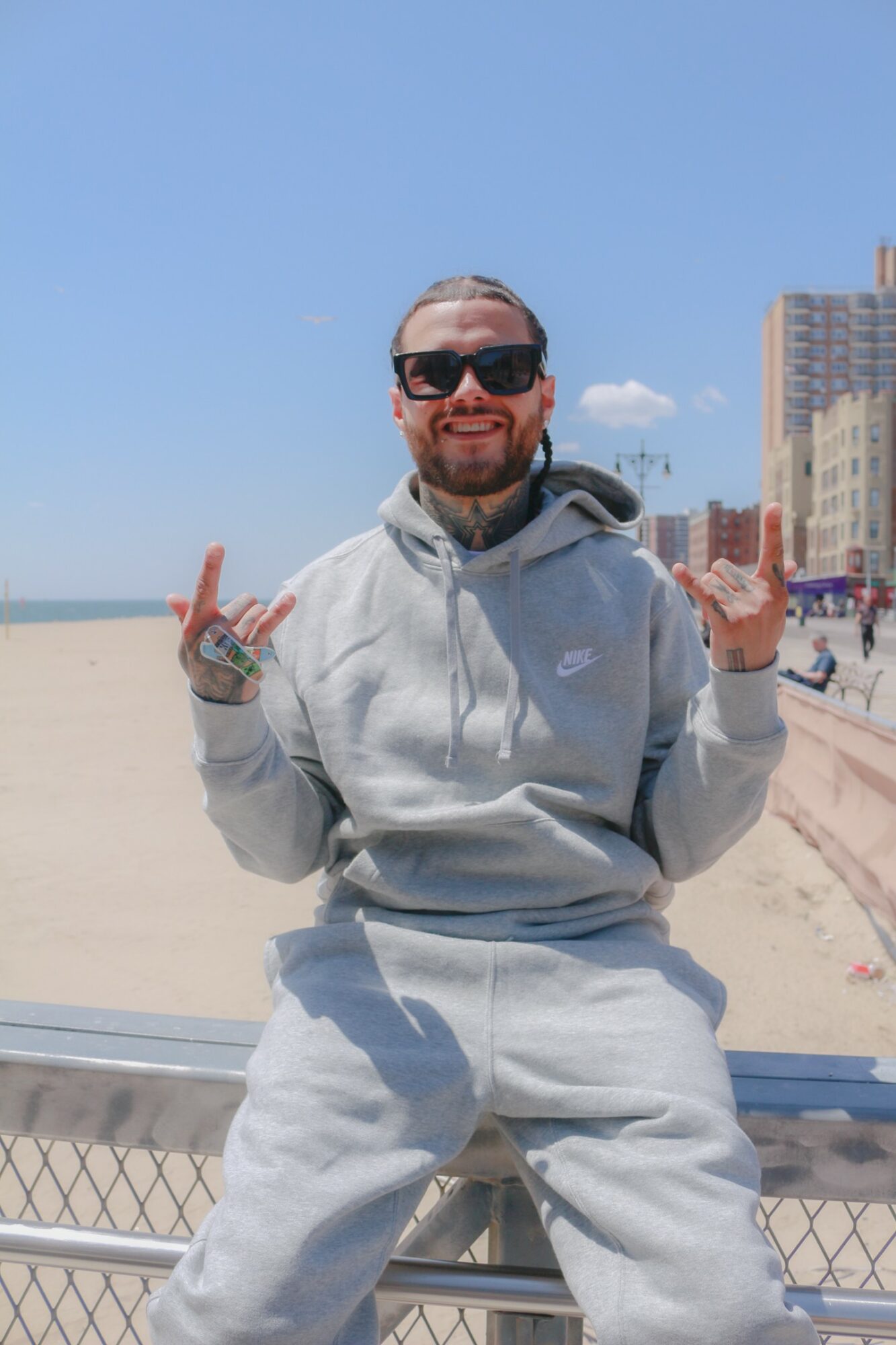 Man in sunglasses and gray hoodie sitting on a railing at the beach, smiling, with buildings in the background.