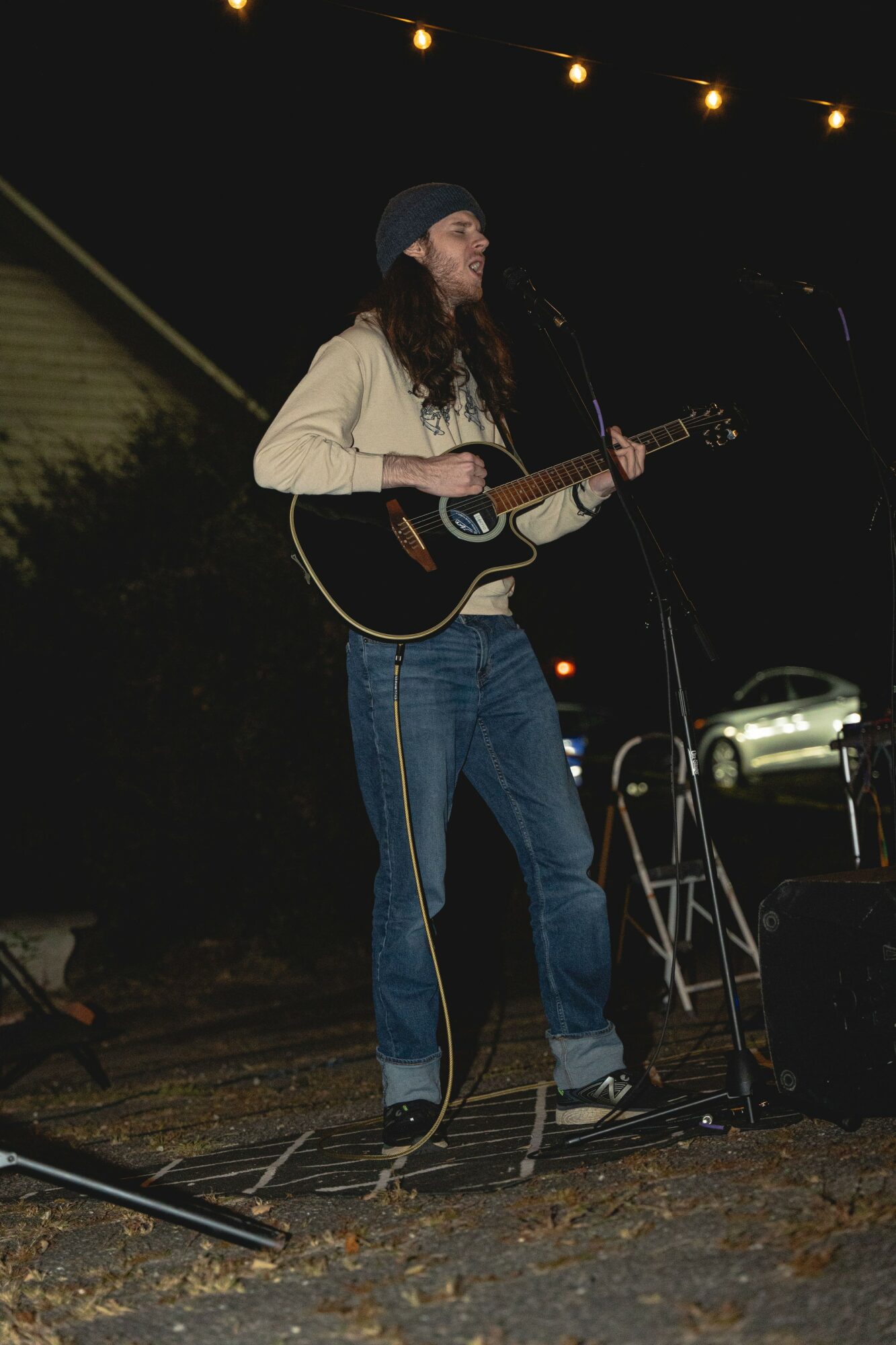 Person with long hair, wearing a beanie, playing guitar and singing on stage at night, with string lights overhead.