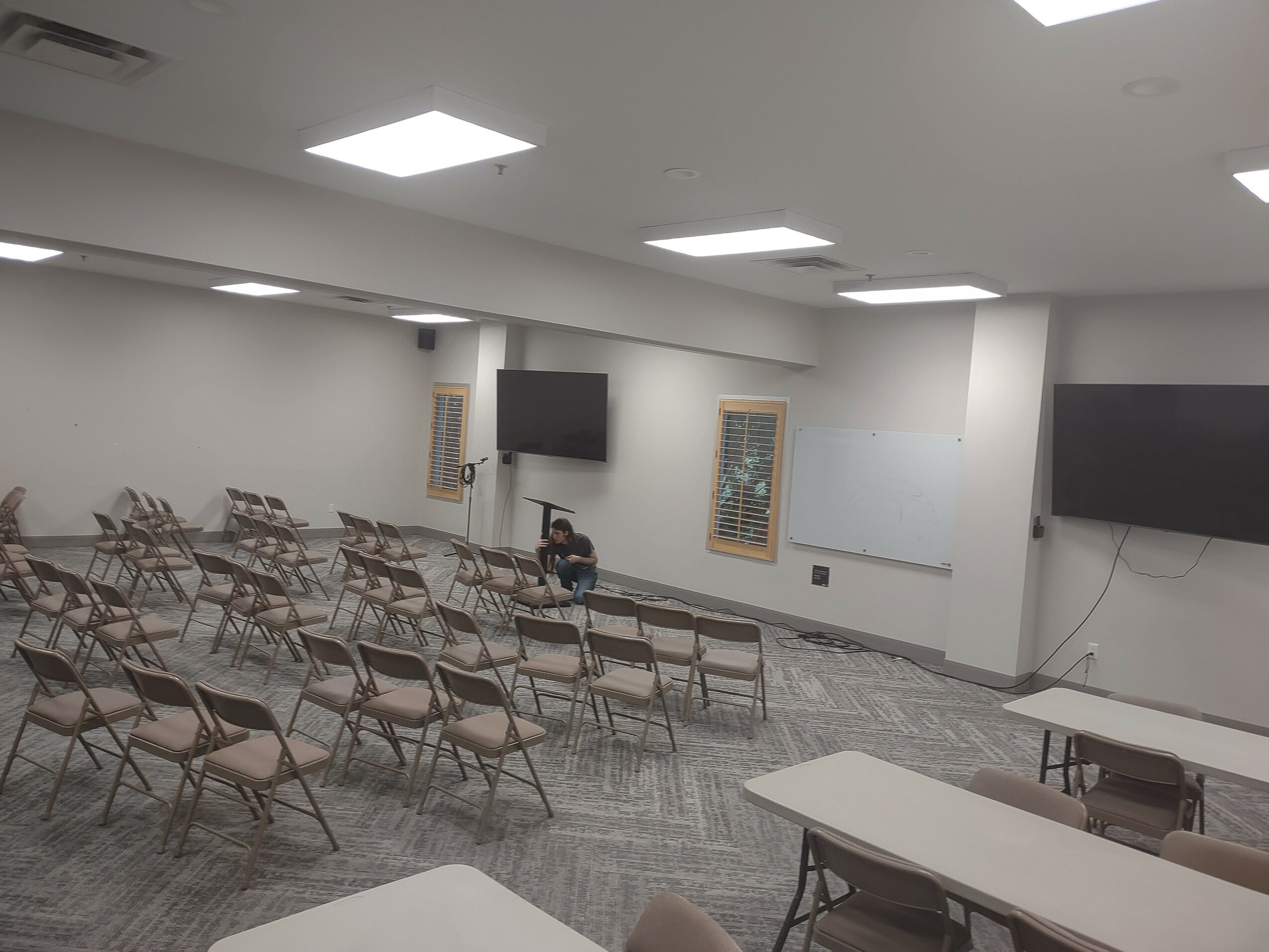 Empty conference room with rows of chairs, two large screens on walls, and a person sitting at the front.