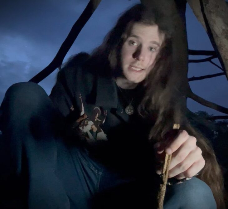 Person with long hair sitting outdoors at dusk, holding a stick, with a tent in the background.