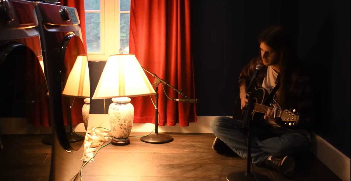Person sitting on the floor playing guitar near a window with red curtains, two lamps, and a dark wall.