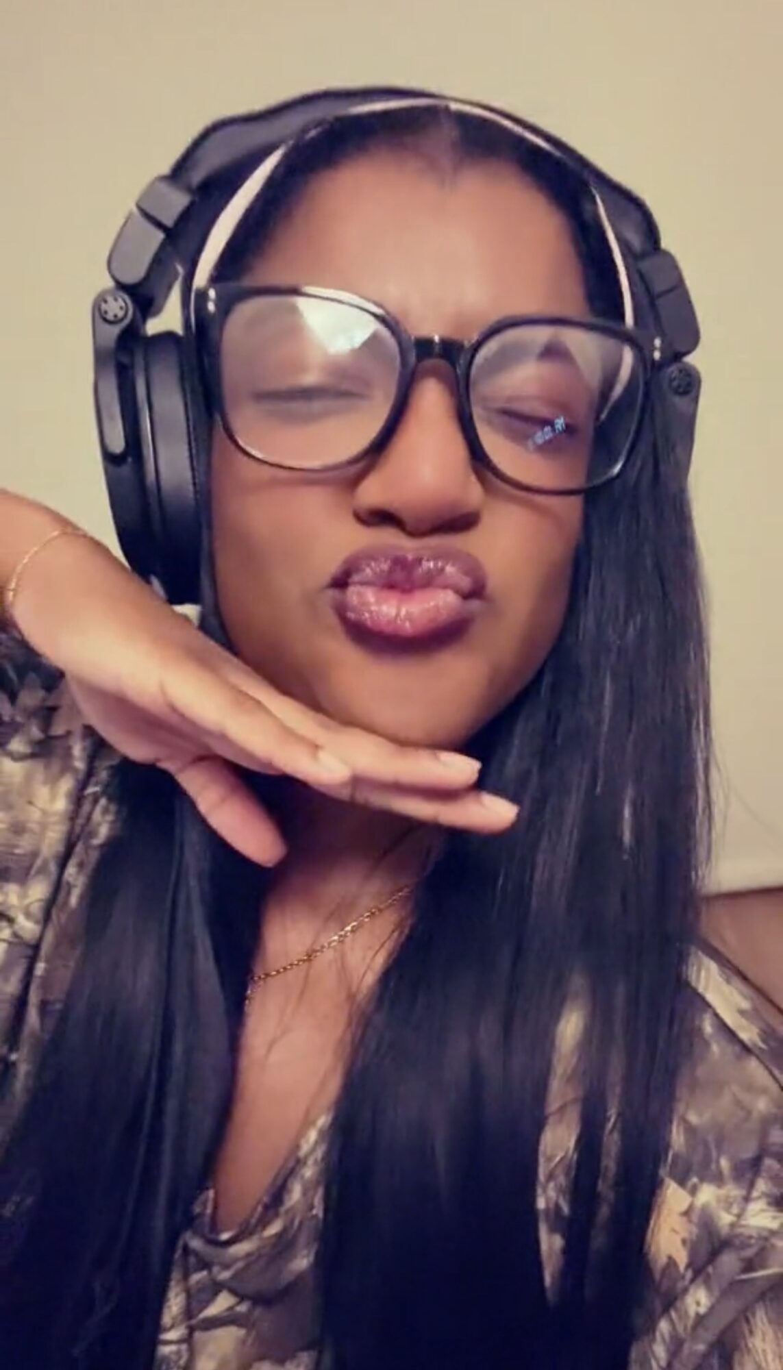 Woman with glasses and headphones making a kiss face, hand under chin, long dark hair, neutral background