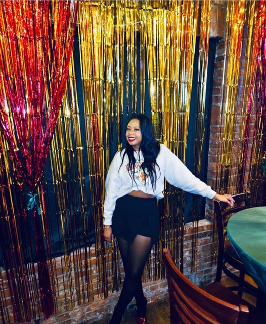 Woman with long dark hair smiling, wearing a white jacket, black skirt, and tights, standing in front of colorful metallic streamers.