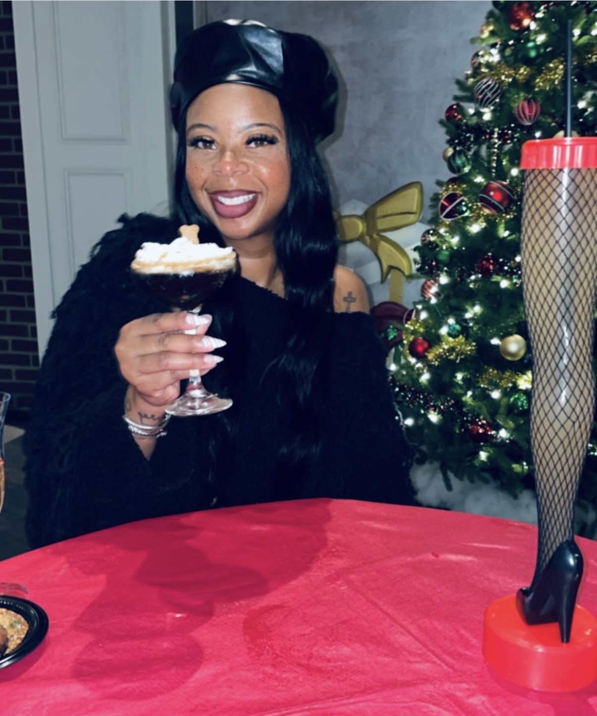 Woman smiling, holding a dessert in a glass, sitting at a table with a red tablecloth, Christmas tree in background.