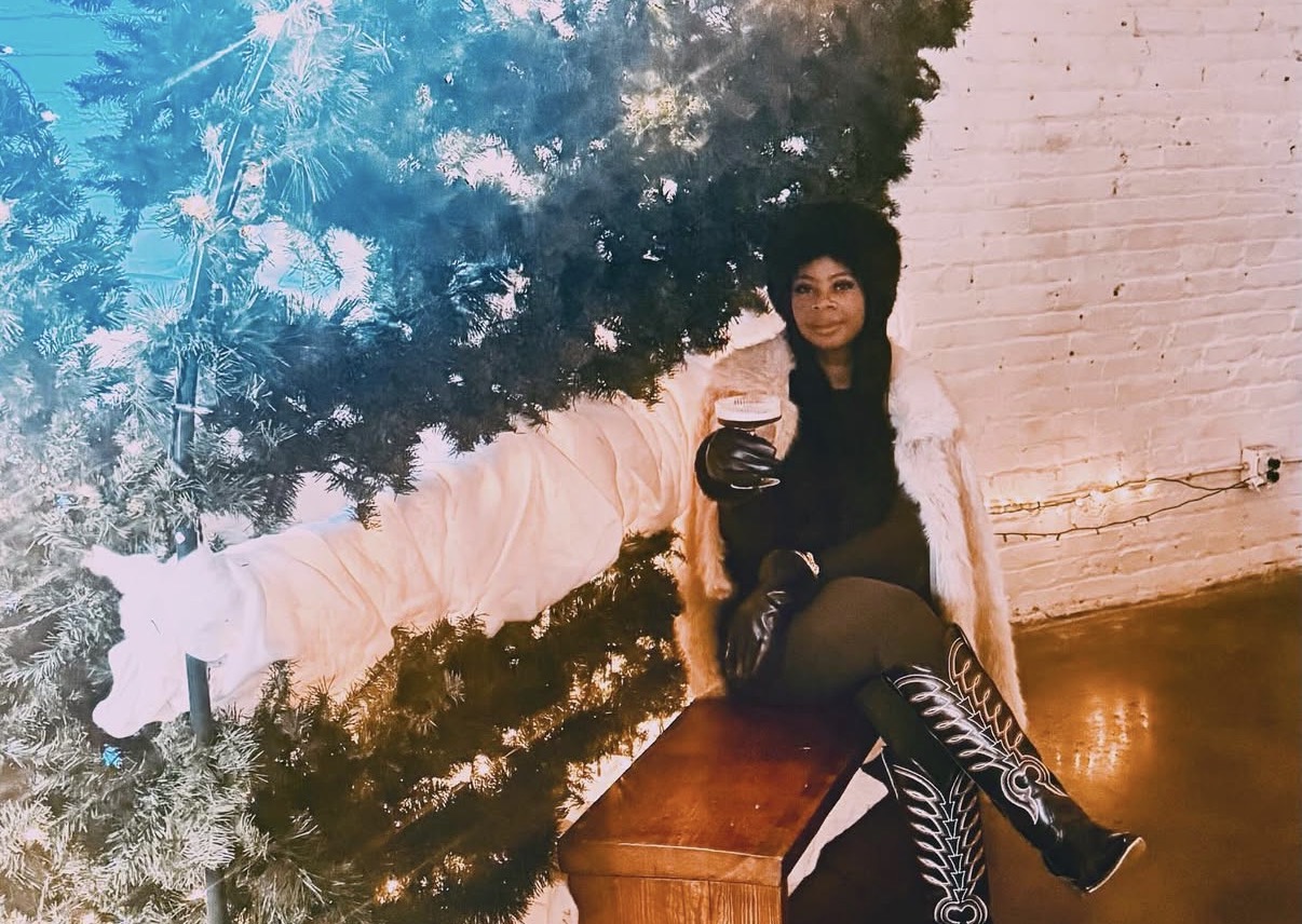 Person sitting on a bench next to a decorated Christmas tree, holding a drink, indoors with a brick wall background.