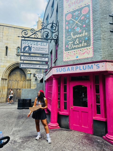 Young girl in black dress and red hat standing outside a pink storefront with a sign reading 'Sugarplum's'.