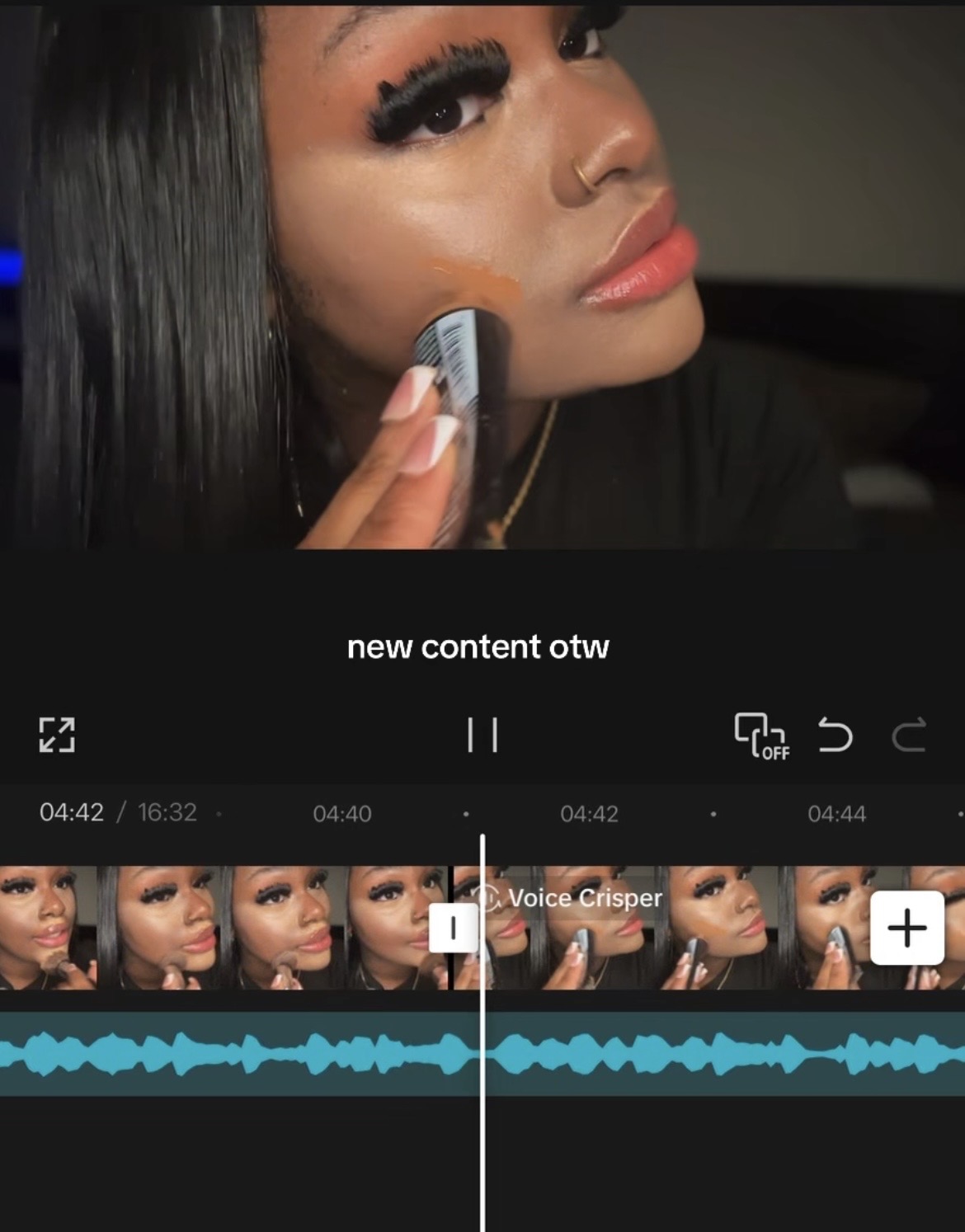Close-up of a woman with long black hair applying makeup with a sponge, looking at the camera, with makeup and earrings visible.