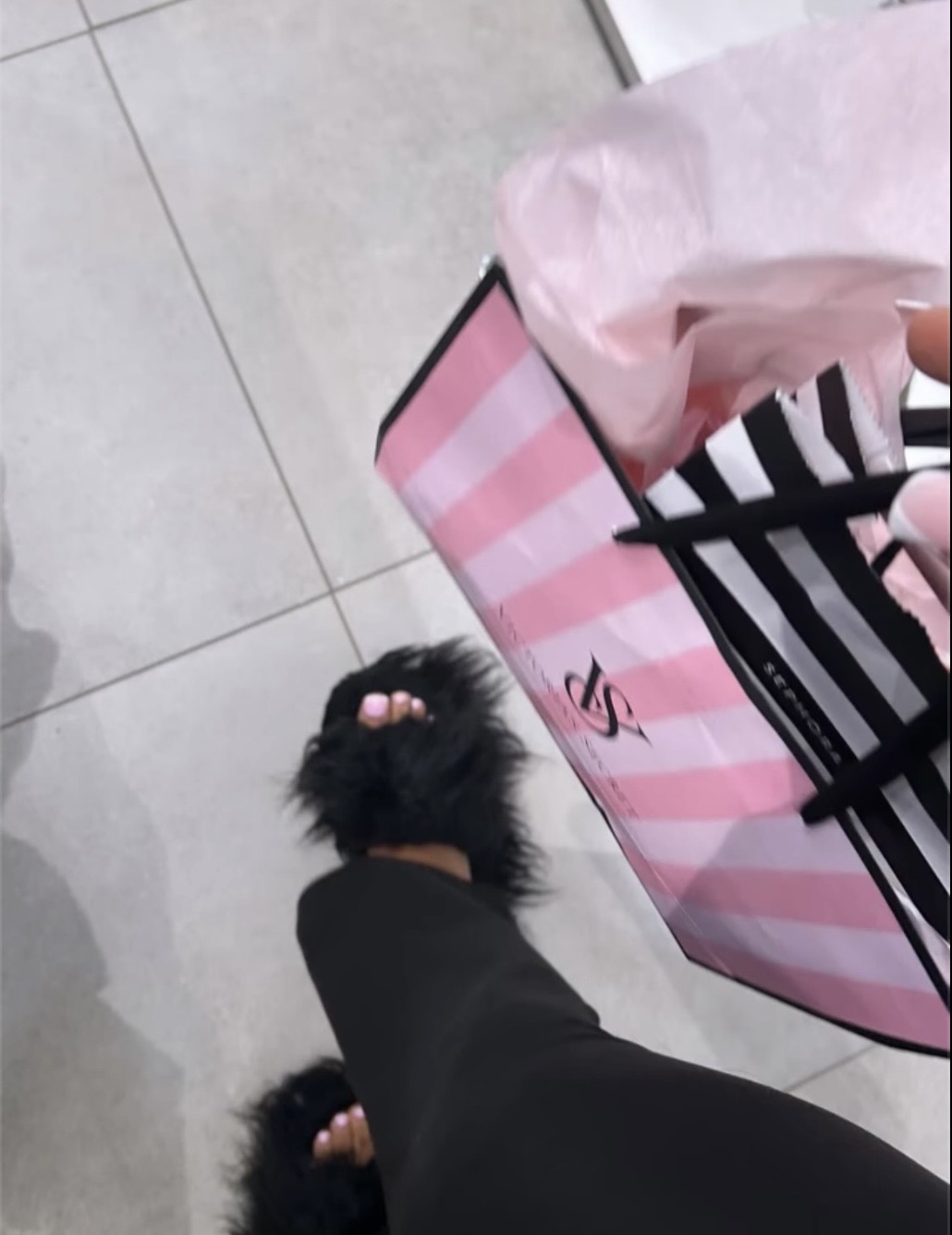 Person's foot wearing a black fluffy slipper on tiled floor, shopping bag and pink tissue paper nearby.