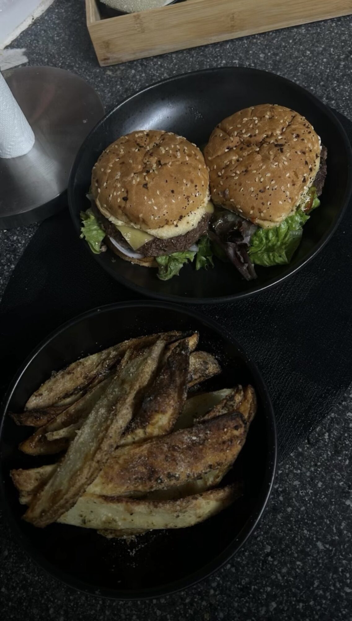 Two burgers with lettuce in a black pan, and a bowl of cooked potato wedges on a dark surface.