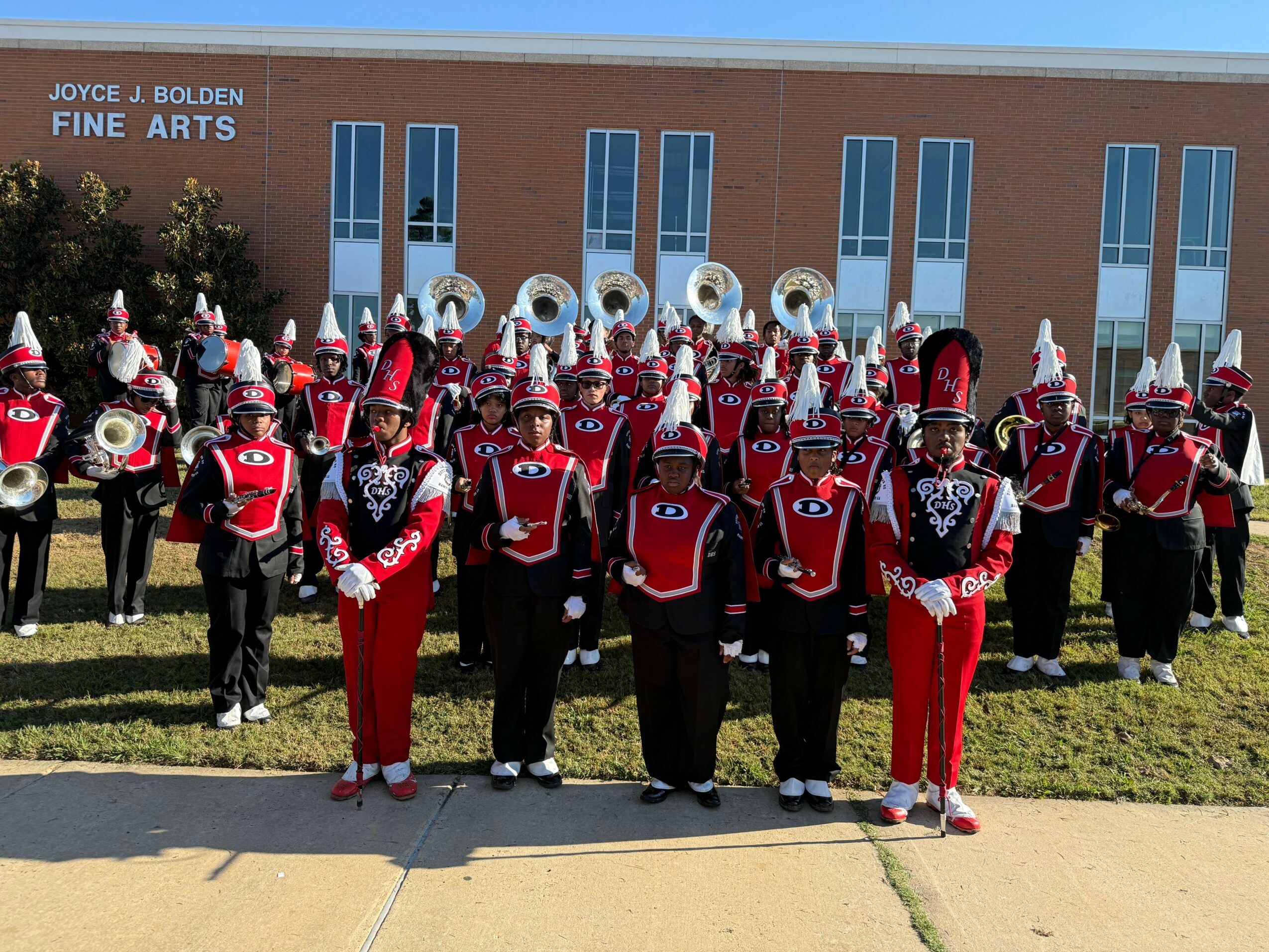 Marching band in red and black uniforms with hats, standing outdoors in front of a brick building with blue windows.