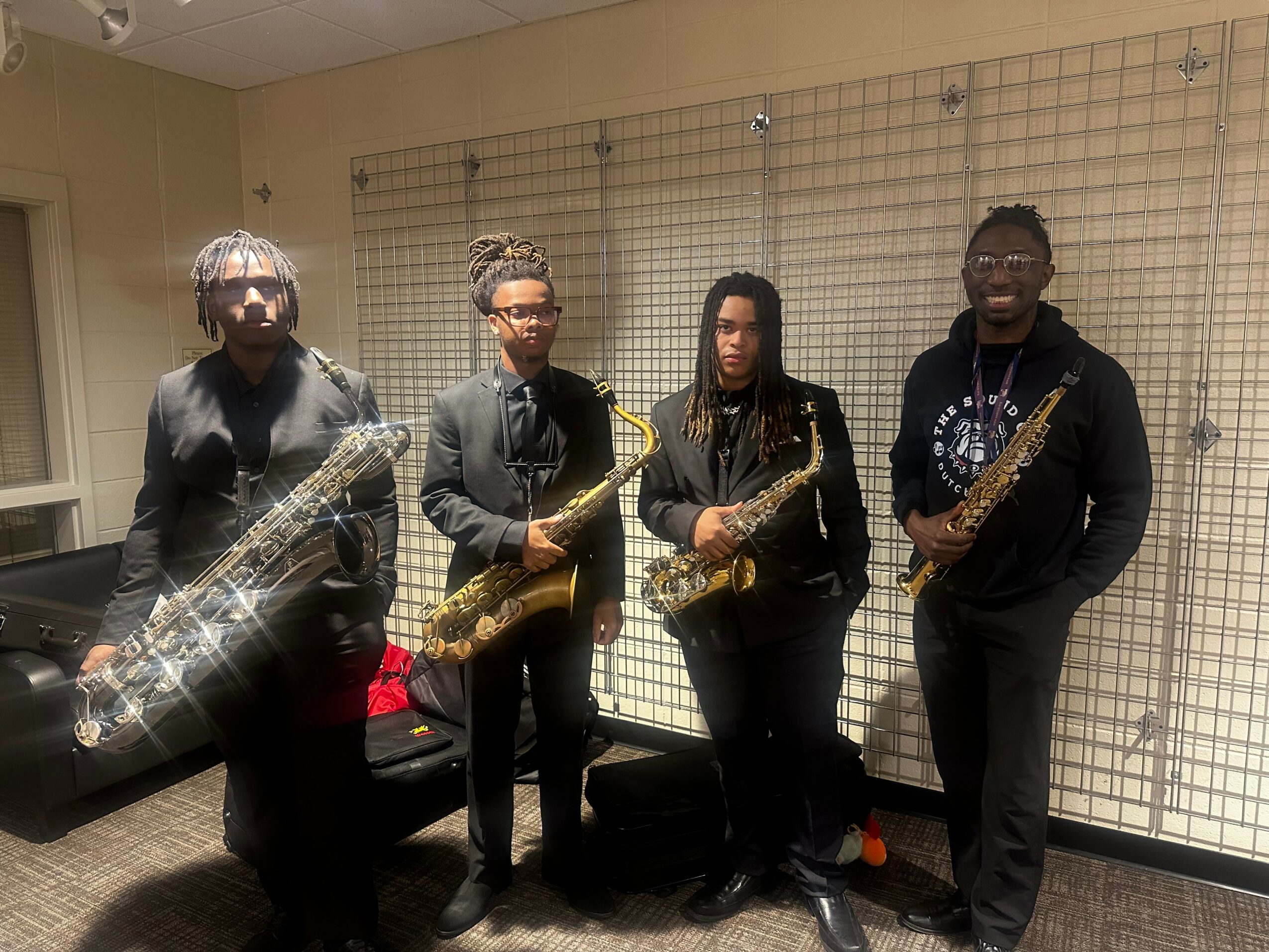 Four people holding saxophones standing indoors in front of a wire grid wall.