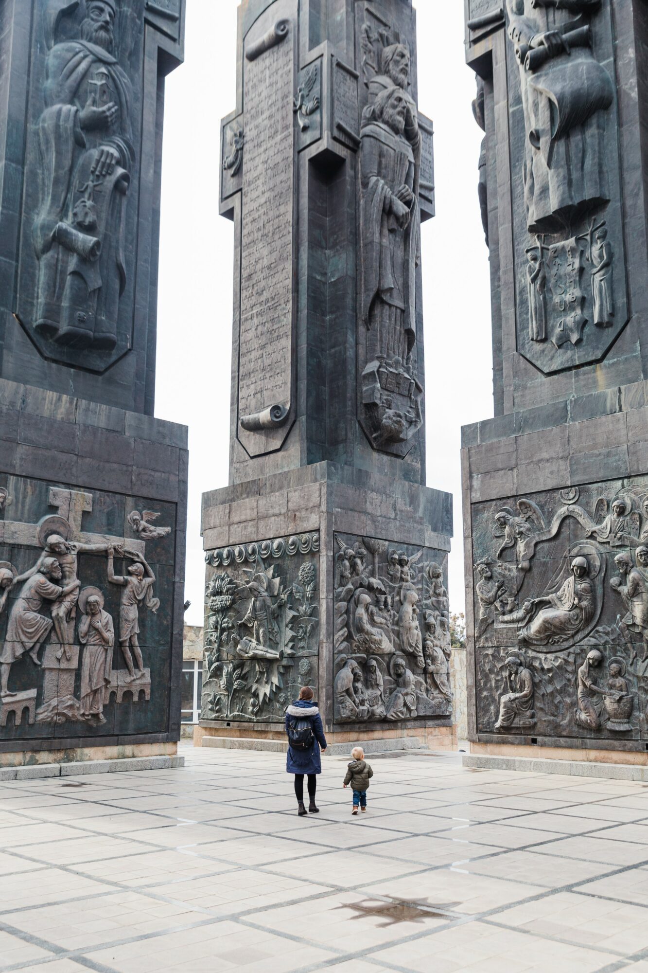 Two people, an adult and a child, stand in front of tall, carved stone monuments with detailed relief sculptures.