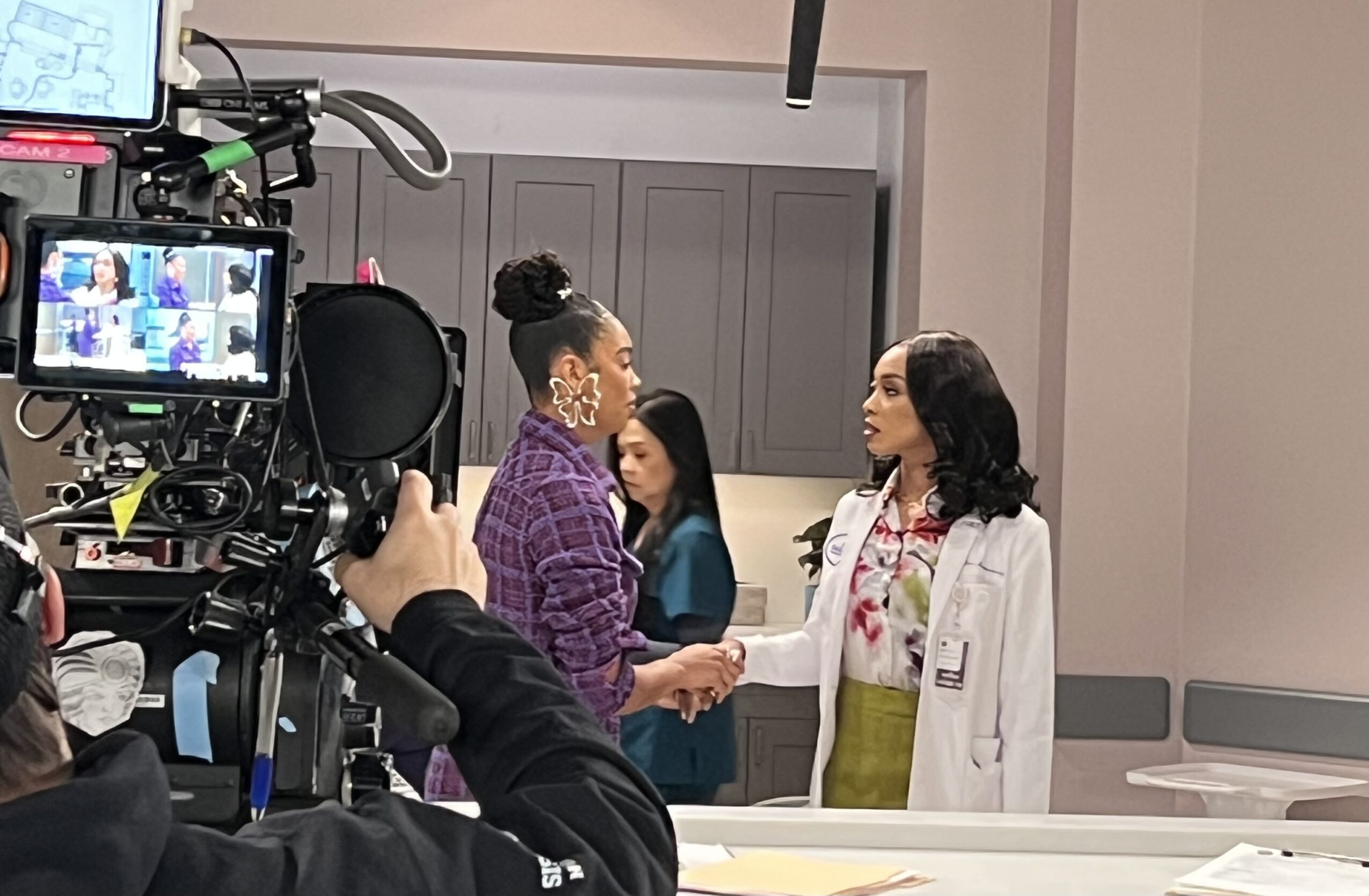 Two women shake hands in a room with a camera filming them, with a third woman in the background. The woman on the right wears a white coat.