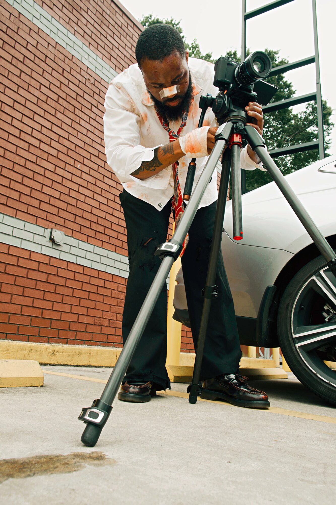 Man operating a camera on a tripod outdoors near a brick wall and a parked car.