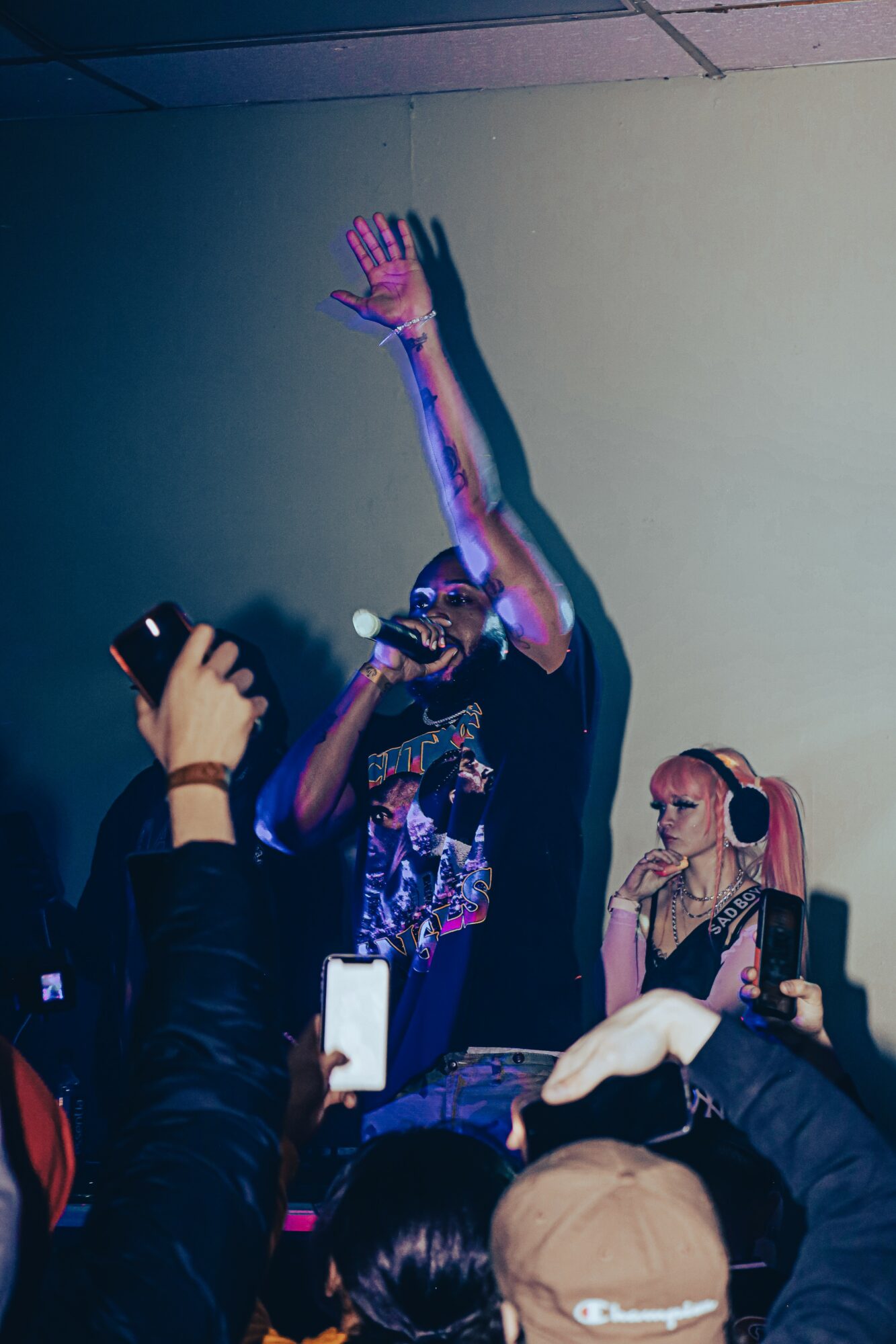 Person singing into microphone with raised hand, surrounded by audience holding phones, in a dimly lit room.