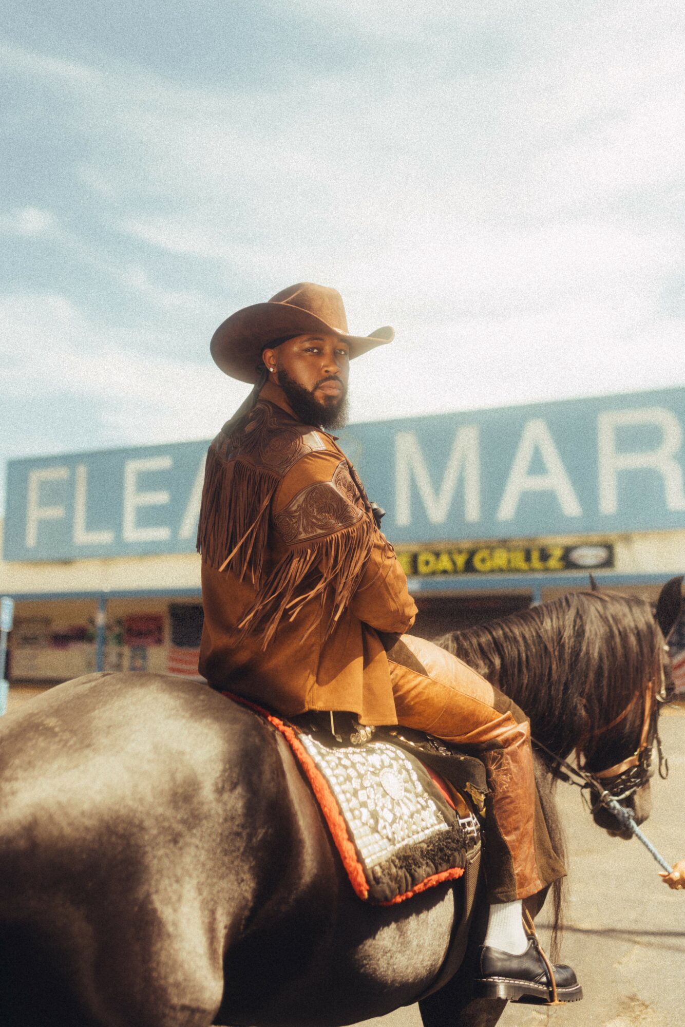 Man wearing a hat and fringed jacket riding a horse in front of a store with a sign reading 'FLEA MARKET'.