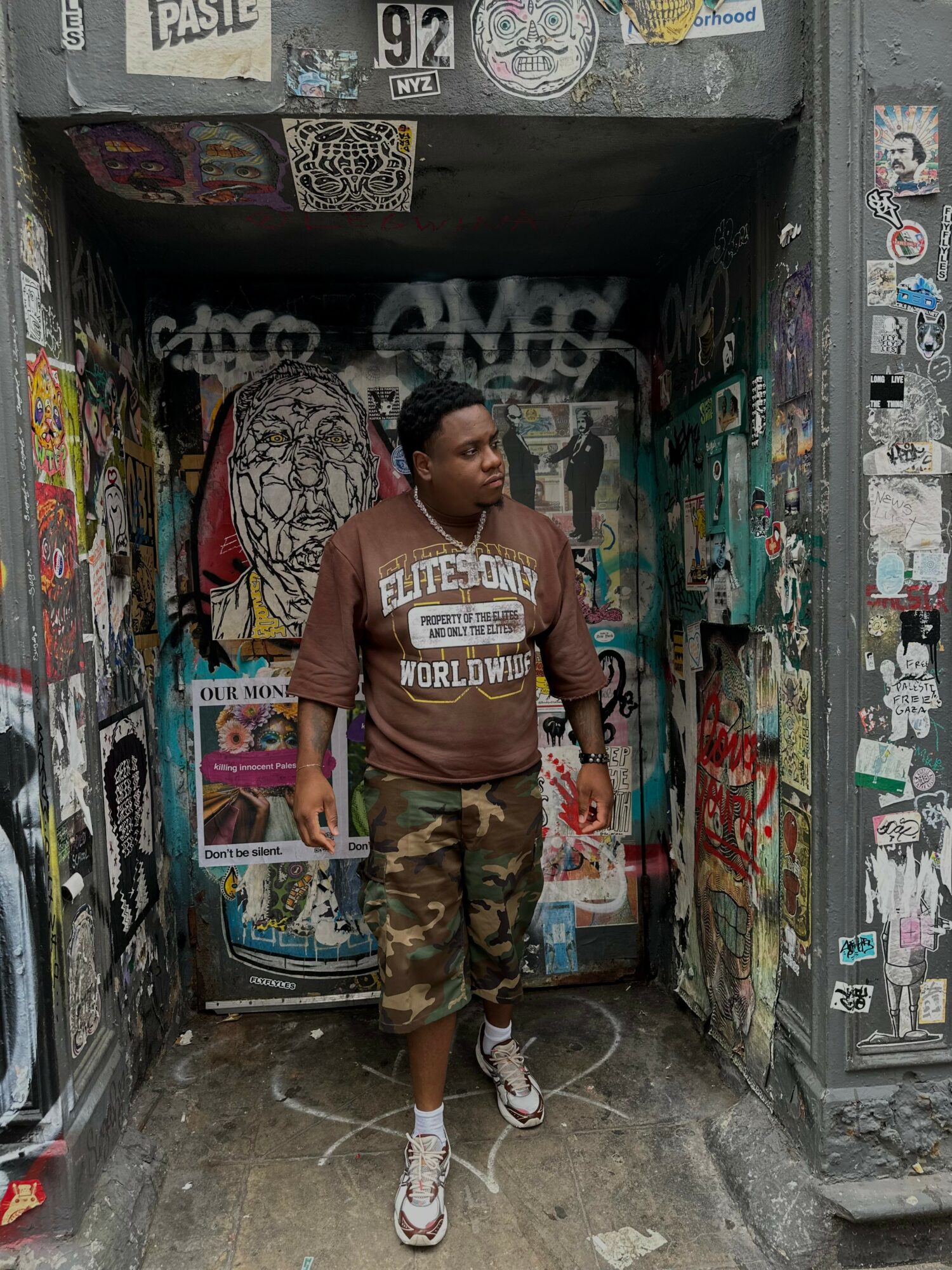 Young person standing in a graffiti-covered doorway, wearing a brown sweatshirt and camouflage shorts, holding a phone.