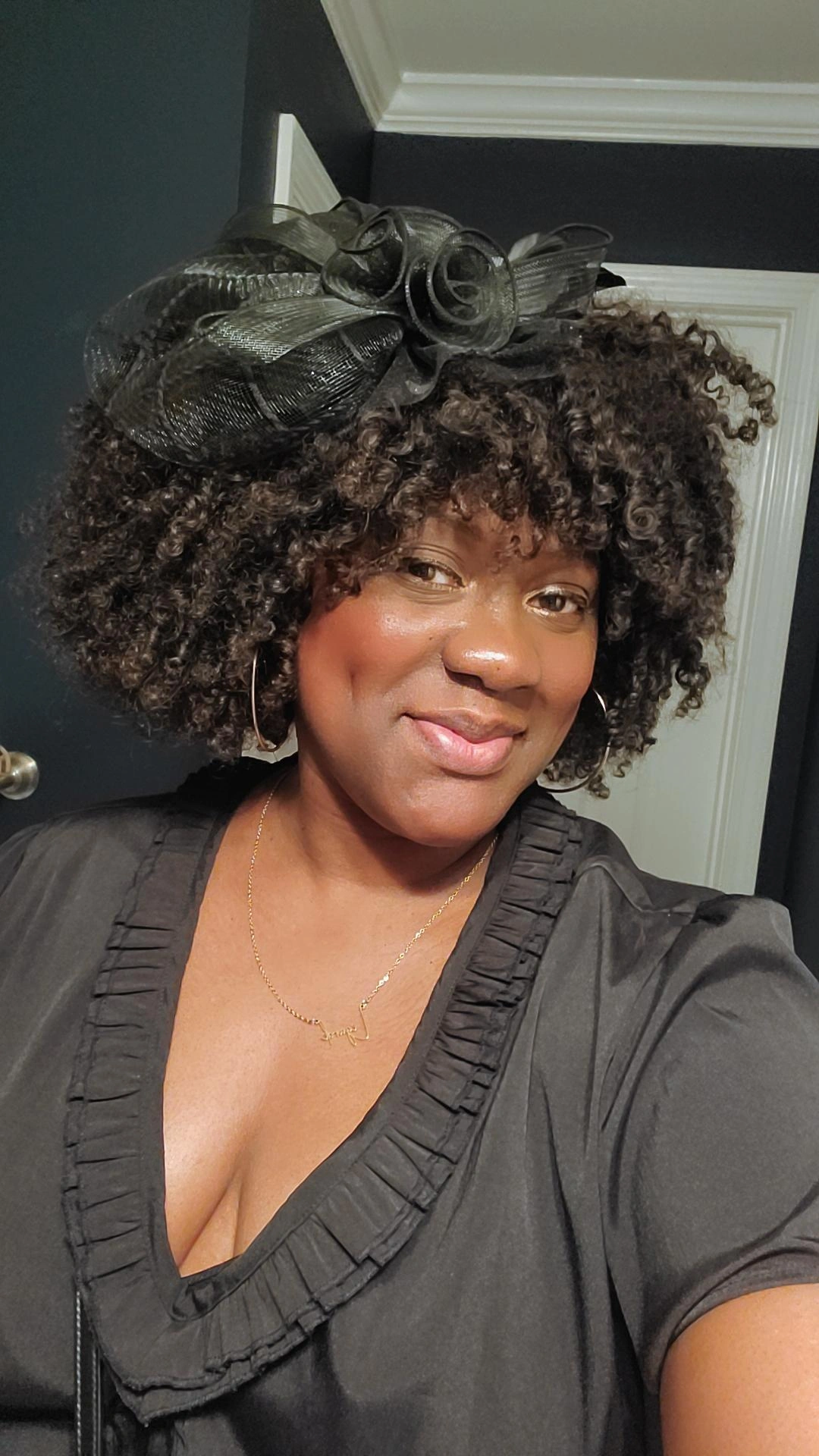Woman with curly hair wearing a black dress and a large black bow headpiece, smiling, indoors.