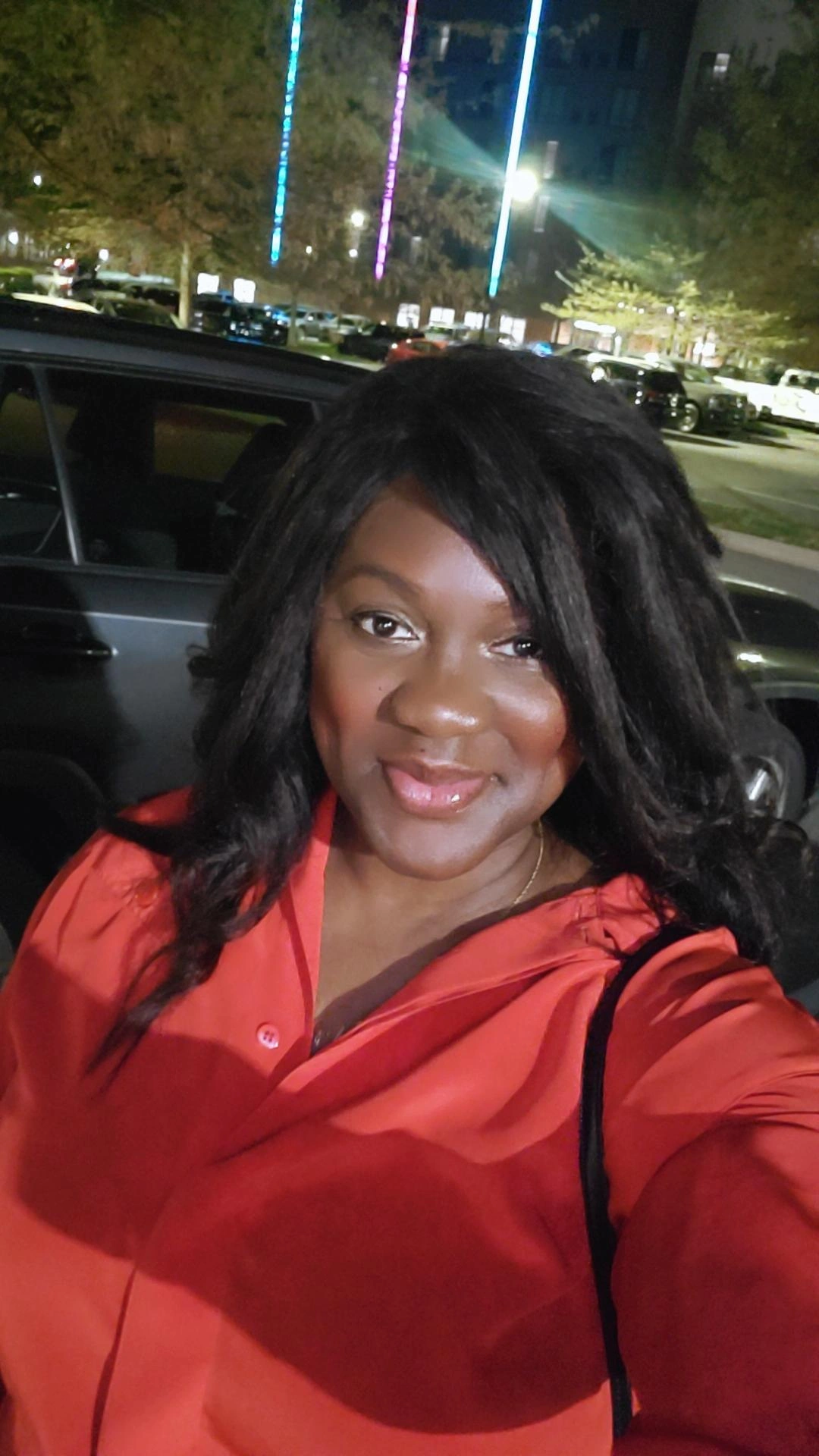 Woman with long black hair wearing a red top, smiling outdoors at night with colorful lights in the background.