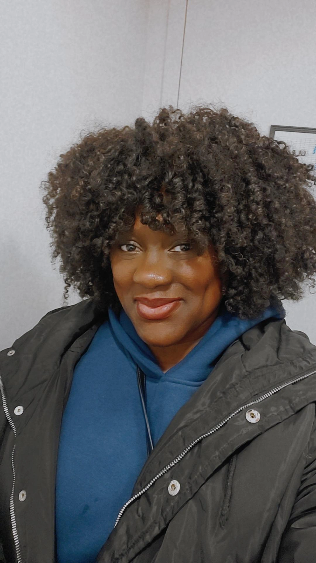 Woman with curly hair smiling, wearing a blue hoodie and black jacket, indoors against a plain wall.