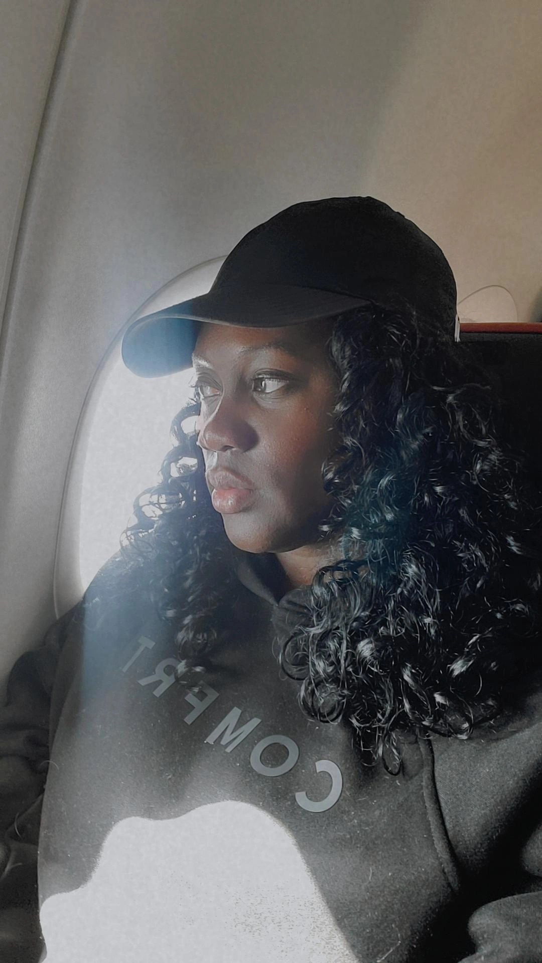 Person with curly hair wearing a cap and hoodie, looking out of an airplane window.