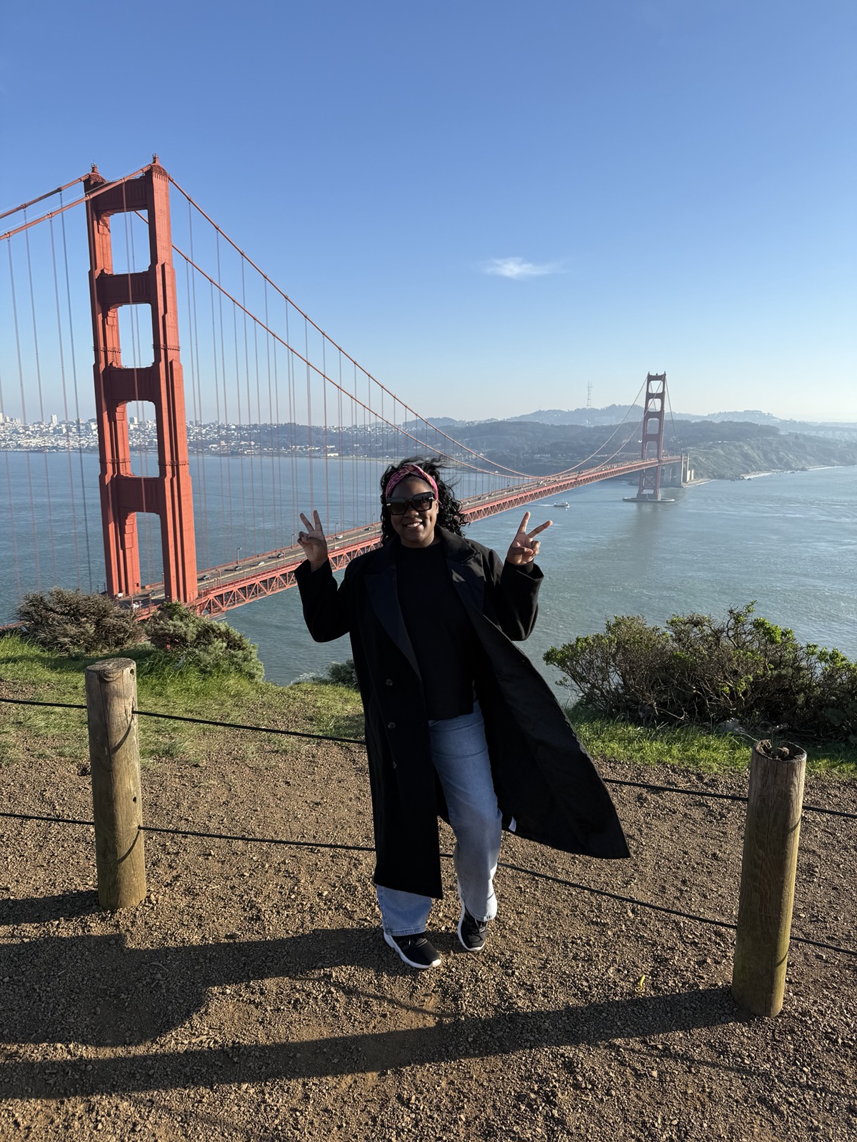 Person standing outdoors near the Golden Gate Bridge, smiling and making peace signs with both hands, with water and hills in the background.