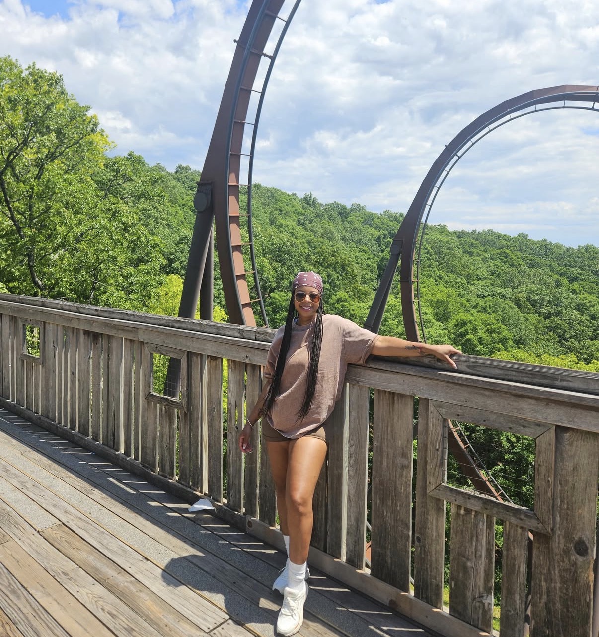 Woman standing on a wooden bridge with roller coaster tracks in the background, wearing sunglasses, a beige outfit, and white boots.
