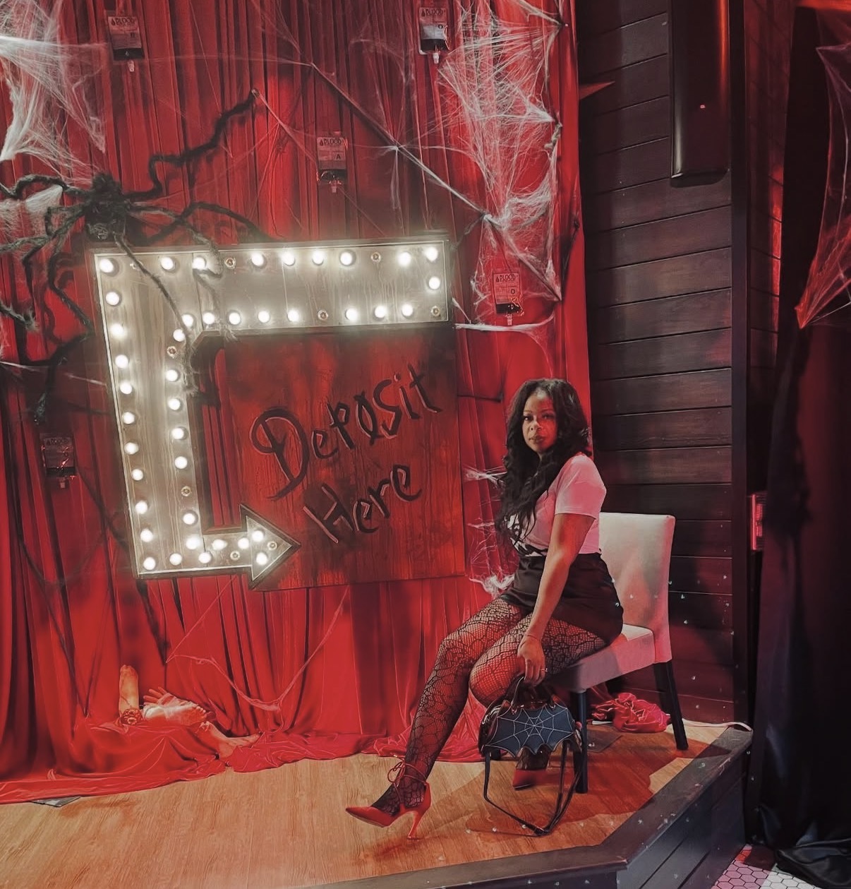 Woman sitting on a chair in front of a Halloween-themed backdrop with spider webs and a large arrow sign, decorated for a spooky event.