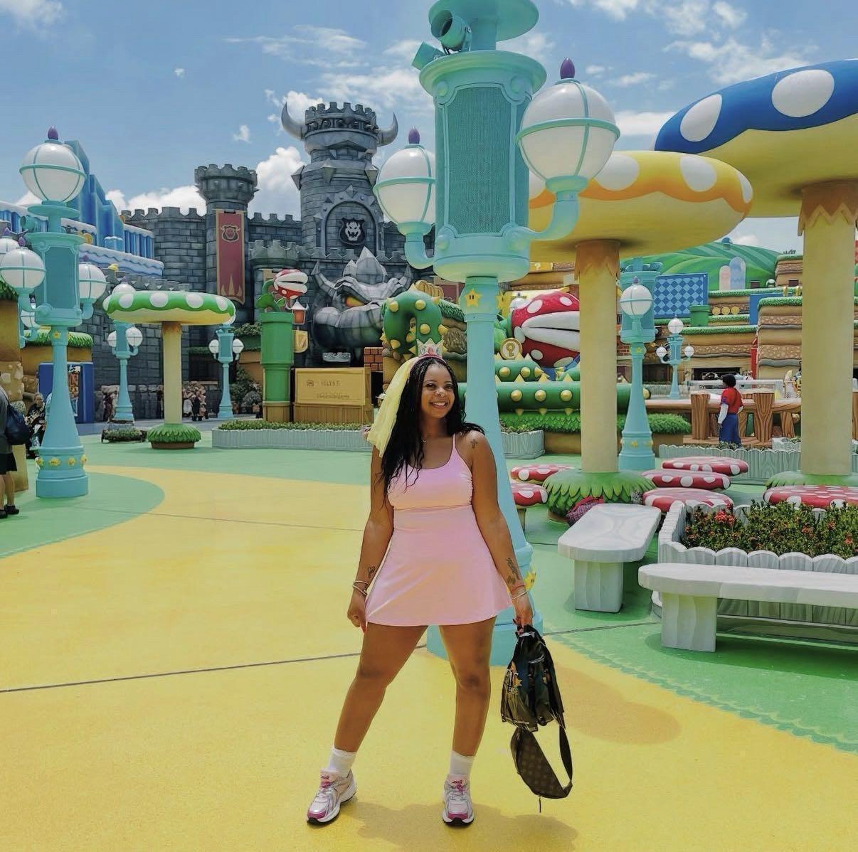 Young woman in pink dress holding a backpack, standing in colorful amusement park with castle and whimsical structures.