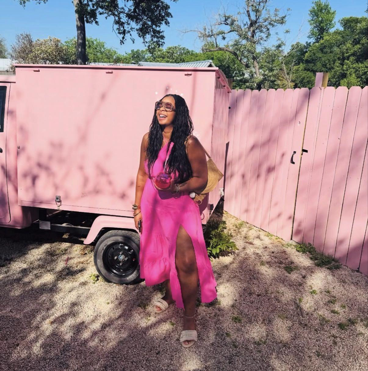 Woman in pink dress with slit, smiling outdoors near pink fence and trailer, holding a drink, sunny day, trees in background.