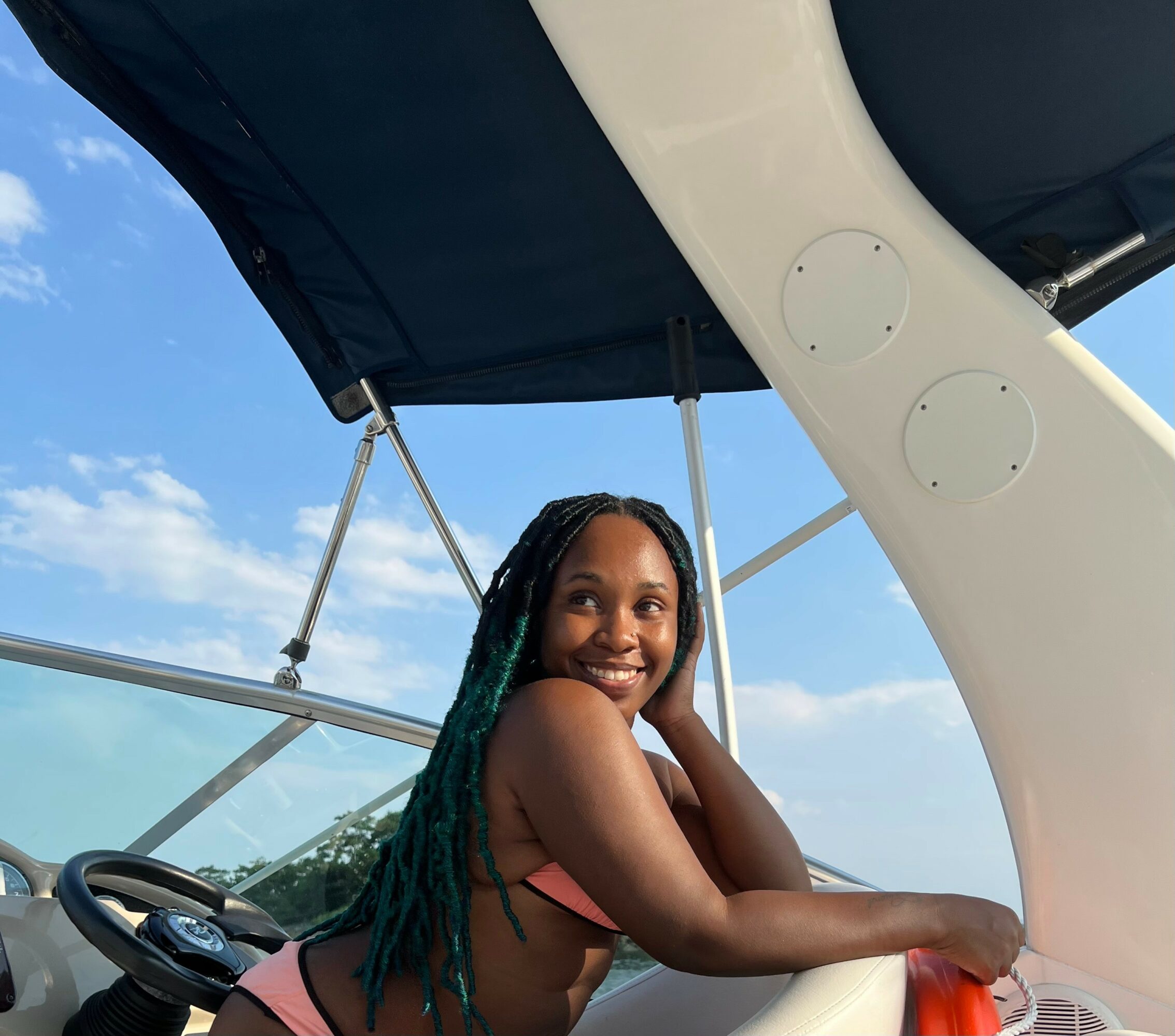 Smiling woman with long braided hair on boat, blue sky with clouds in background.